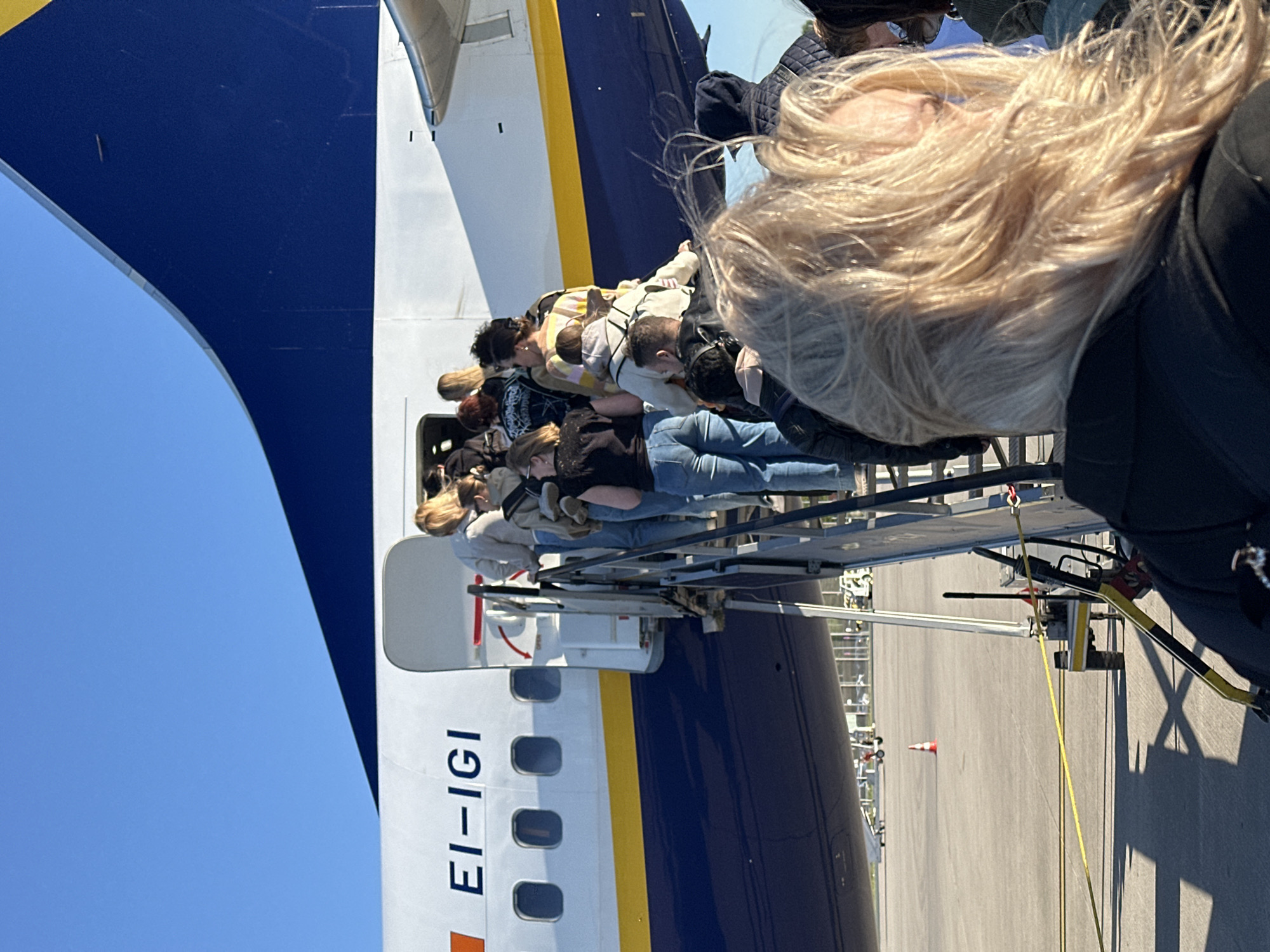 The image depicts passengers boarding a commercial aircraft at an airport, likely in Weeze, Germany. The perspective is from behind a person with long blonde hair, who is in the immediate foreground, looking forward towards the aircraft. A line of other passengers is visible ahead, ascending a mobile boarding staircase to enter the aircraft's fuselage.
The aircraft is a distinctive dark blue and white, separated by a yellow stripe running horizontally along its side. The registration number "EI-IGI" is clearly visible in large black letters on the white part of the fuselage, identifying it as a Ryanair Boeing 737. The boarding stairs are metal, connecting the tarmac to the aircraft door.
The scene takes place under bright, clear blue skies, indicating good weather conditions during the daytime. The tarmac can be seen below the aircraft and stairs, with a small orange cone faintly visible in the distance on the right side of the tarmac. The overall impression is a routine and busy aircraft boarding process.