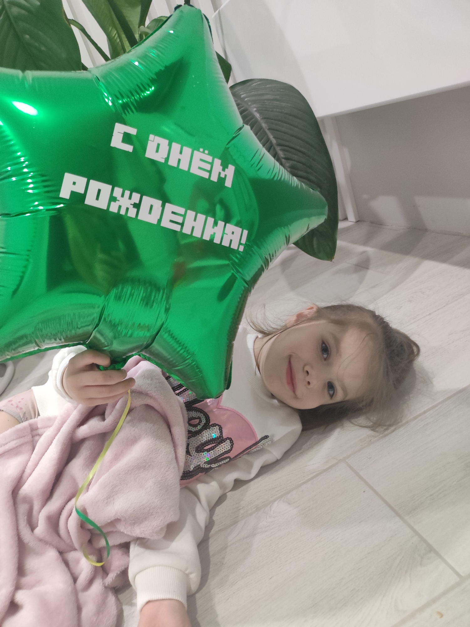 A young girl lies on a light-colored, wood-patterned floor, holding a large, green, star-shaped balloon with the Cyrillic inscription "С Днём Рождения!" (Happy Birthday!). She is wearing a white sweatshirt with pink accents and sequins. A light pink blanket partially covers her. The scene is indoors, near a plant in a pot and a white wall. The location is presumed to be within a building in Votkinsk, Russia.