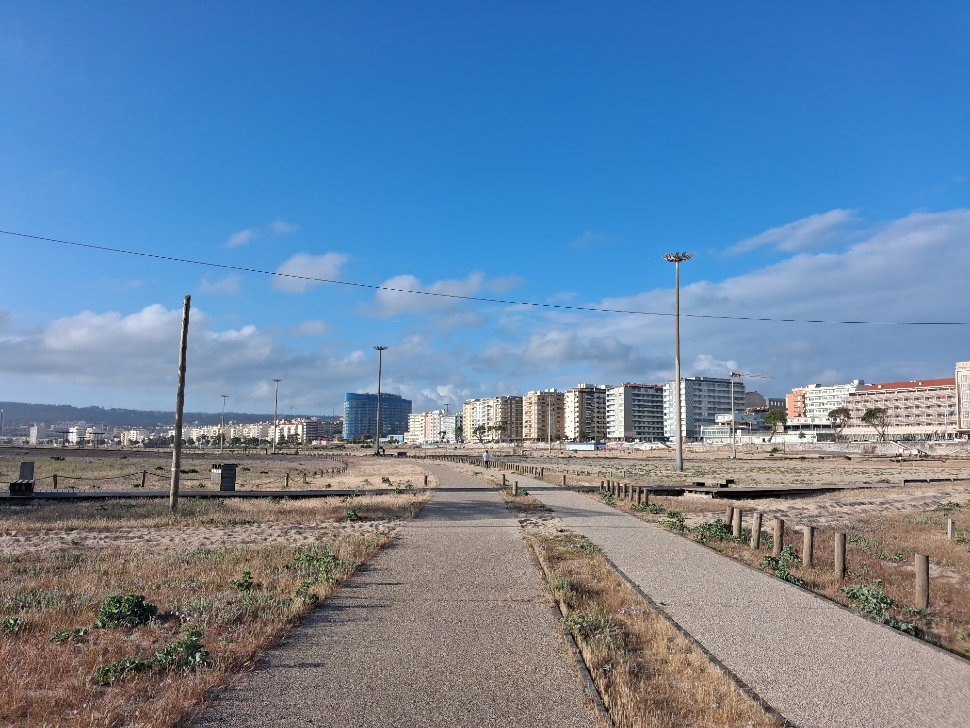 The image shows a view in Figueira da Foz, Portugal, looking towards a line of buildings from a pedestrian walkway that stretches into the distance. The walkway is flanked by grassy, sandy areas with sparse vegetation and wooden posts. The sky is blue with scattered clouds. Overhead power lines run across the frame. Several streetlights are visible along the path. In the background, a variety of multi-story buildings can be seen, ranging in color and design. Construction equipment is present on the top of one of the buildings. The day appears to be sunny.