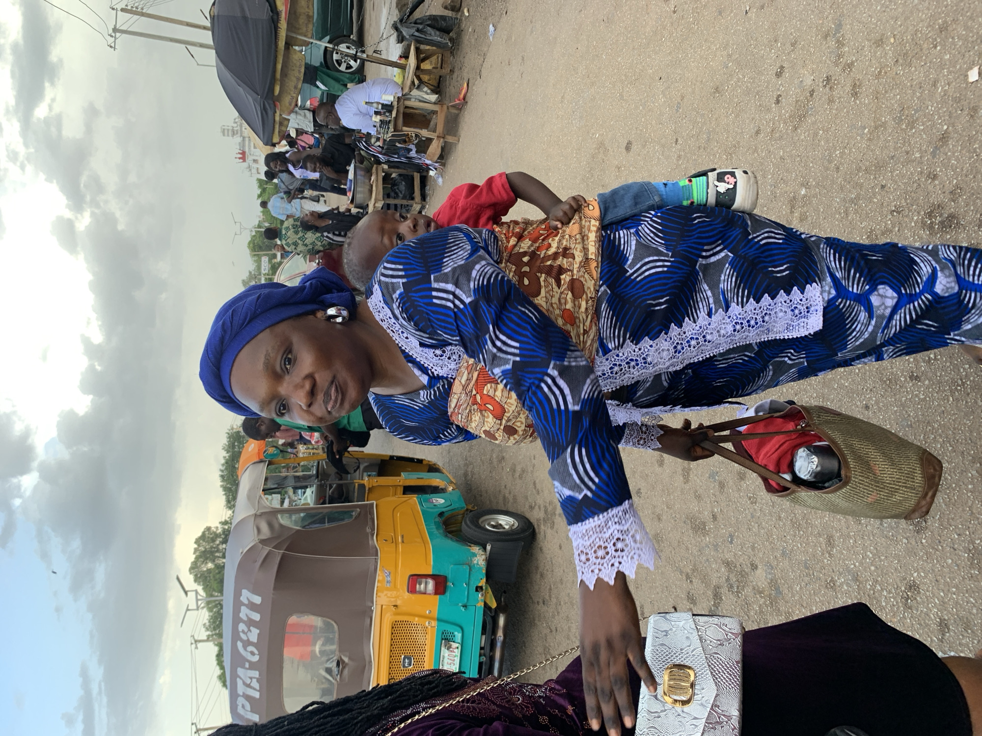 The image captures a street scene in Jos, Nigeria, under a cloudy sky. In the foreground, a woman wearing a blue headwrap and a blue and white patterned dress is carrying a young child on her back, secured with a patterned fabric wrap. The child is wearing a red shirt and blue jeans. The woman is holding a woven bag containing some items. To the left, another person, partially visible, is wearing a dark purple dress and holding a light-colored textured handbag. Behind them, a yellow and turquoise tricycle, identified by the text "PTA-6277" on its canopy, is parked. In the background, several other people are visible, some interacting near small stalls and a black umbrella. Cars and utility poles are also present, contributing to the urban environment. The ground appears to be a dirt or gravel road.