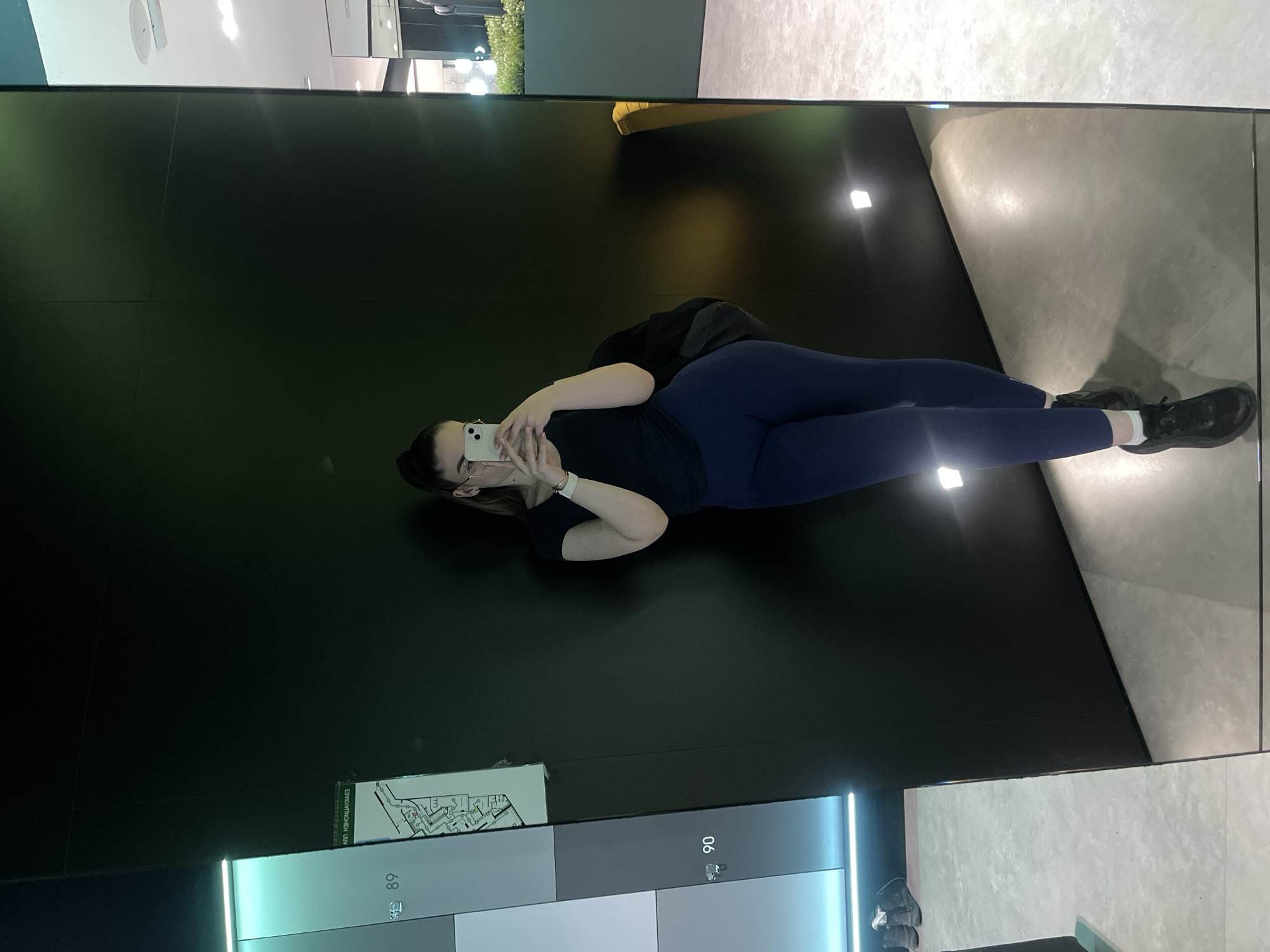 A woman is lying down taking a photo in a reflective black room, likely a store or showroom. She is wearing a black top, dark blue leggings, and black shoes with white socks. She's holding a white smartphone, presumably taking a selfie. The environment features a black wall, a light gray tiled floor reflected in the wall, and signage at the bottom that includes floor plan diagrams. The location appears to be indoors, but specific city details are not discernible from the image.