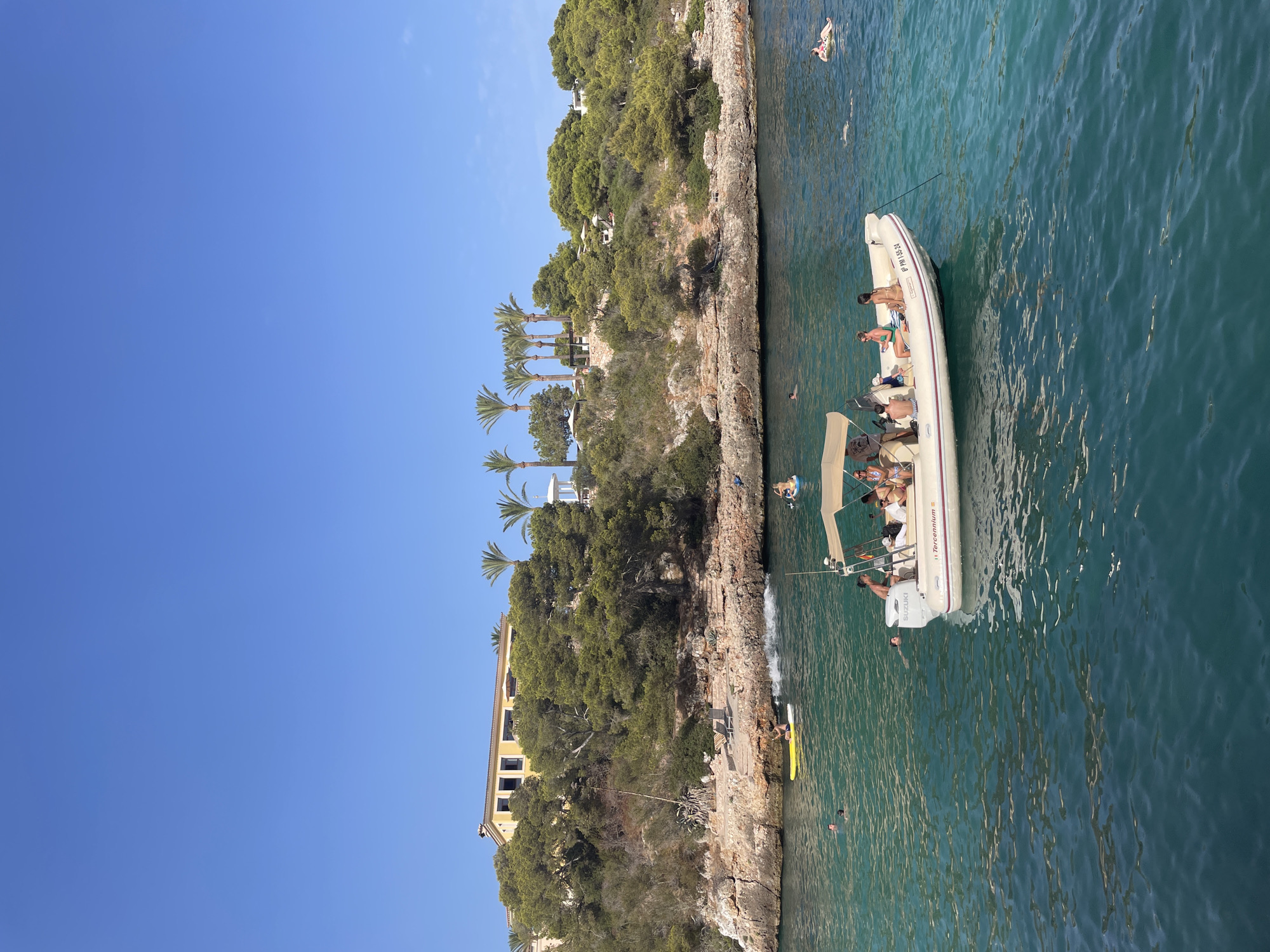 An aerial view shows a bright blue sky above a coastal landscape in Cala Ferrera, Spain. The turquoise waters of the bay are calm, with ripples distorting the underwater landscape. A small white boat with a beige canopy is anchored in the foreground, carrying six people, some of whom are sitting and others standing. The boat has the registration "6° PM 1 55-24" on its stern. In the middle ground, several people are swimming and floating in the water, with two individuals on paddleboards. One paddleboarder is creating a splash as they move. The background features a rocky, tree-covered hillside with a large yellow building situated on its crest. Palm trees are visible behind the building, along with a white gazebo. The rocky coastline is dotted with dense green vegetation, and stone steps lead down to the water.