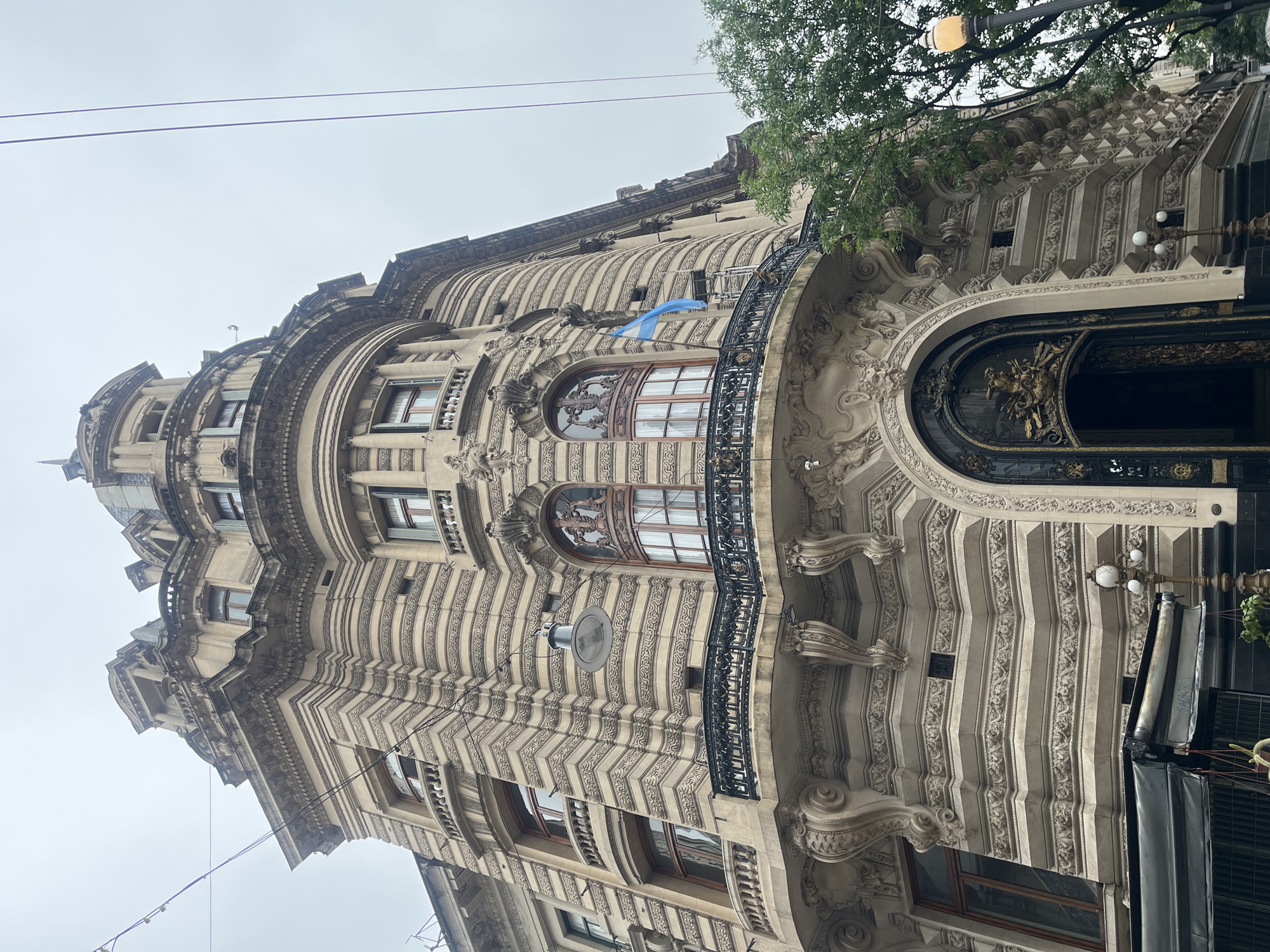 The image shows a building in Buenos Aires, Argentina. The building's architecture is ornate, with detailed carvings, a balcony, and rounded windows. There is an Argentinian flag hanging from the building. The sky is overcast. A tree branch and street lamp are visible at the top right corner of the image.