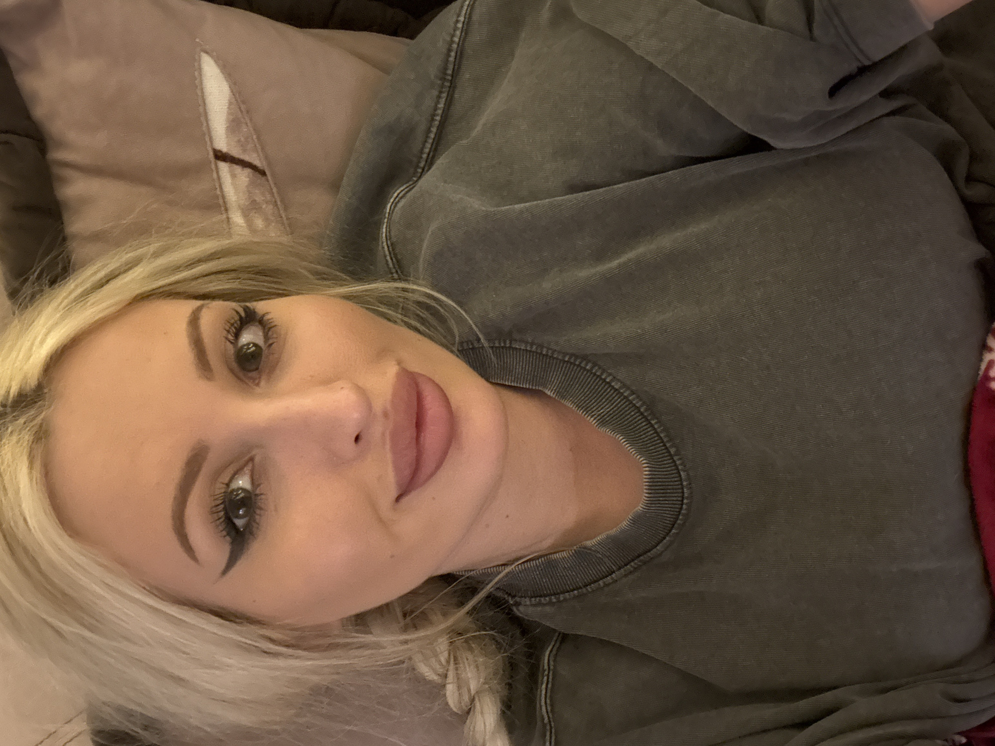 A woman with blonde hair, heavy makeup, and a grey sweatshirt is lying on a couch. The woman is looking at the camera and smiling. The background is a blurred couch with a pillow. The lighting is dim.