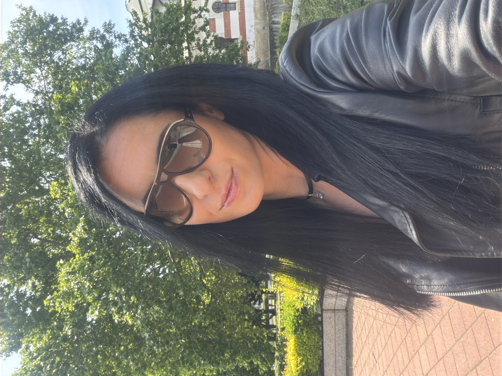 A woman is posing for a photo. She is wearing a black leather jacket and sunglasses. The background shows trees and a building. The location is Pleven, Bulgaria, in front of the Pleven Panorama.