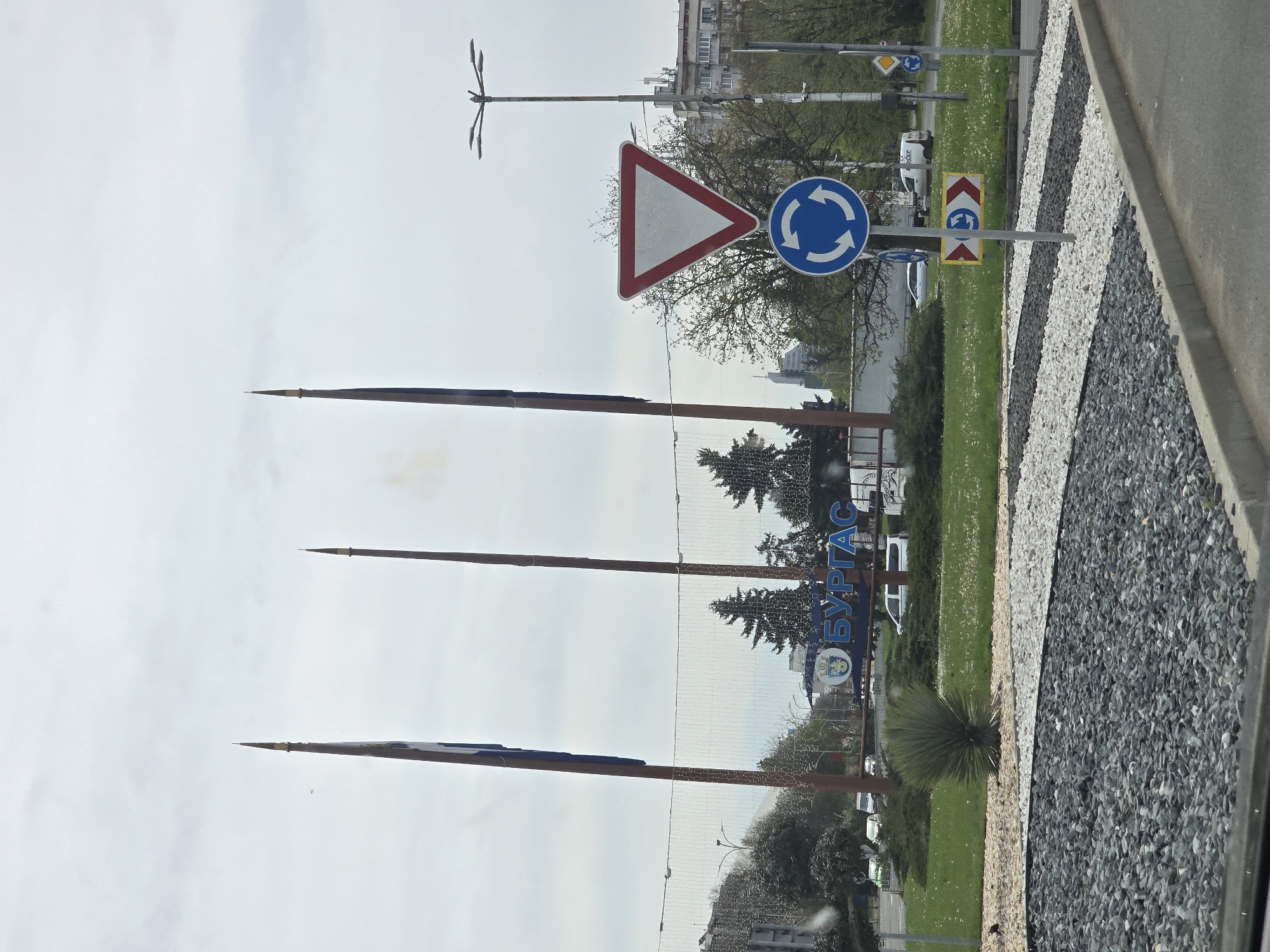 The image, captured in a vertical orientation, displays a daytime roadside scene at what appears to be an entrance or major intersection in Burgas, Bulgaria. Three prominent, tall, slender, spire-like metal structures, possibly decorative monuments, rise vertically against a light gray, overcast sky.
In the foreground, a decorative median or island features alternating stripes of dark gray and light beige/white crushed stone. Adjacent to this, a grassy area is visible. A paved road runs along the right edge of the frame.
Several European-style road signs are prominently displayed. A red-bordered white triangular sign, indicating "Give Way" or "Yield," is positioned next to a blue circular sign with white arrows, signifying a "Roundabout." Further along, additional road signs include another blue "Roundabout" sign, a yellow diamond-shaped "Priority Road" sign, and a red and yellow chevron sign typically indicating an obstacle or a specific direction for a roundabout. A street light with multiple arms extends from the left.
In the mid-ground, a large blue sign with white Cyrillic text clearly reads "БУРГАС" (Burgas), confirming the location. The area features green landscaping with various trees, including evergreens, and a distinctive spiky yucca-like shrub. Buildings and faint outlines of vehicles are visible in the background, partially obscured by the trees and general haze of an overcast day. No specific activity or people are visible in the static scene. The weather appears to be cloudy, without direct sunlight.