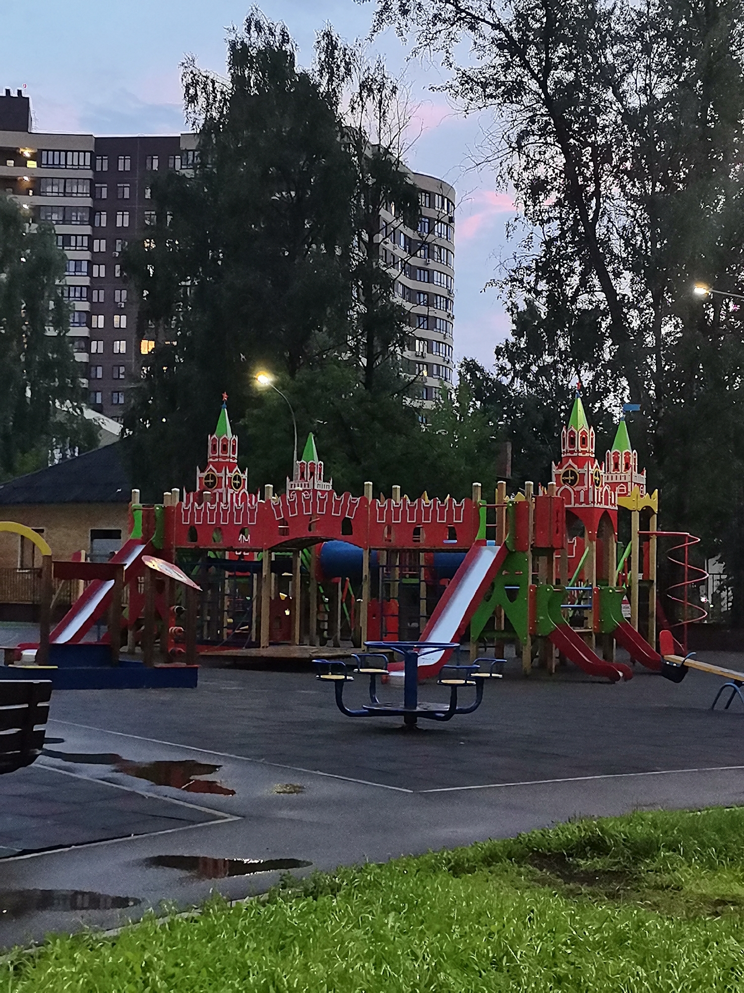The image depicts a children's playground in Krasnogorsk, Russia, at dusk. The playground features a large, multi-colored structure resembling a castle with red walls, green turrets topped with flags, and various slides, climbing areas, and platforms. In the background, two tall residential buildings with illuminated windows are visible, along with dense foliage from mature trees that partially obscure the buildings. The sky shows hints of twilight with a soft pink hue. The playground is situated on an asphalt surface, with several puddles reflecting the surroundings, suggesting recent rain. A blue picnic table with seats and a seesaw are also present on the playground. A portion of a dark-colored bench is visible in the lower left corner. The overall scene is serene, with no people present.