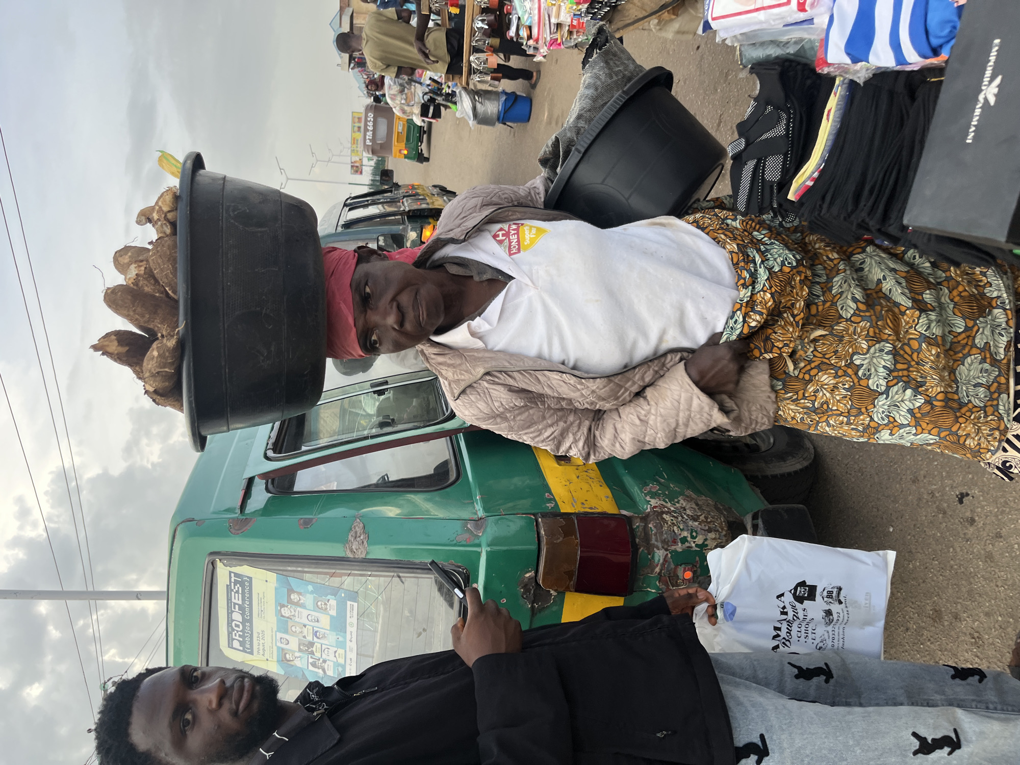 In Bukuru, Nigeria, a woman balances a large black basin filled with yams on her head while standing beside a green bus. She wears a red headscarf, a quilted light brown jacket over a white polo shirt with a "HONEY W" logo, and a colorful patterned skirt. Next to her, a young man in a black jacket and light-wash jeans with black cat logos looks towards the camera, holding a smartphone. The bus, adorned with a "PRO FEST" poster featuring portraits, shows signs of wear and tear, including a yellow stripe and damaged rear lights. The background reveals a street scene with other vehicles, including a three-wheeled auto-rickshaw with the license plate "PTA-6650," and market stalls displaying various goods. The sky is overcast.
