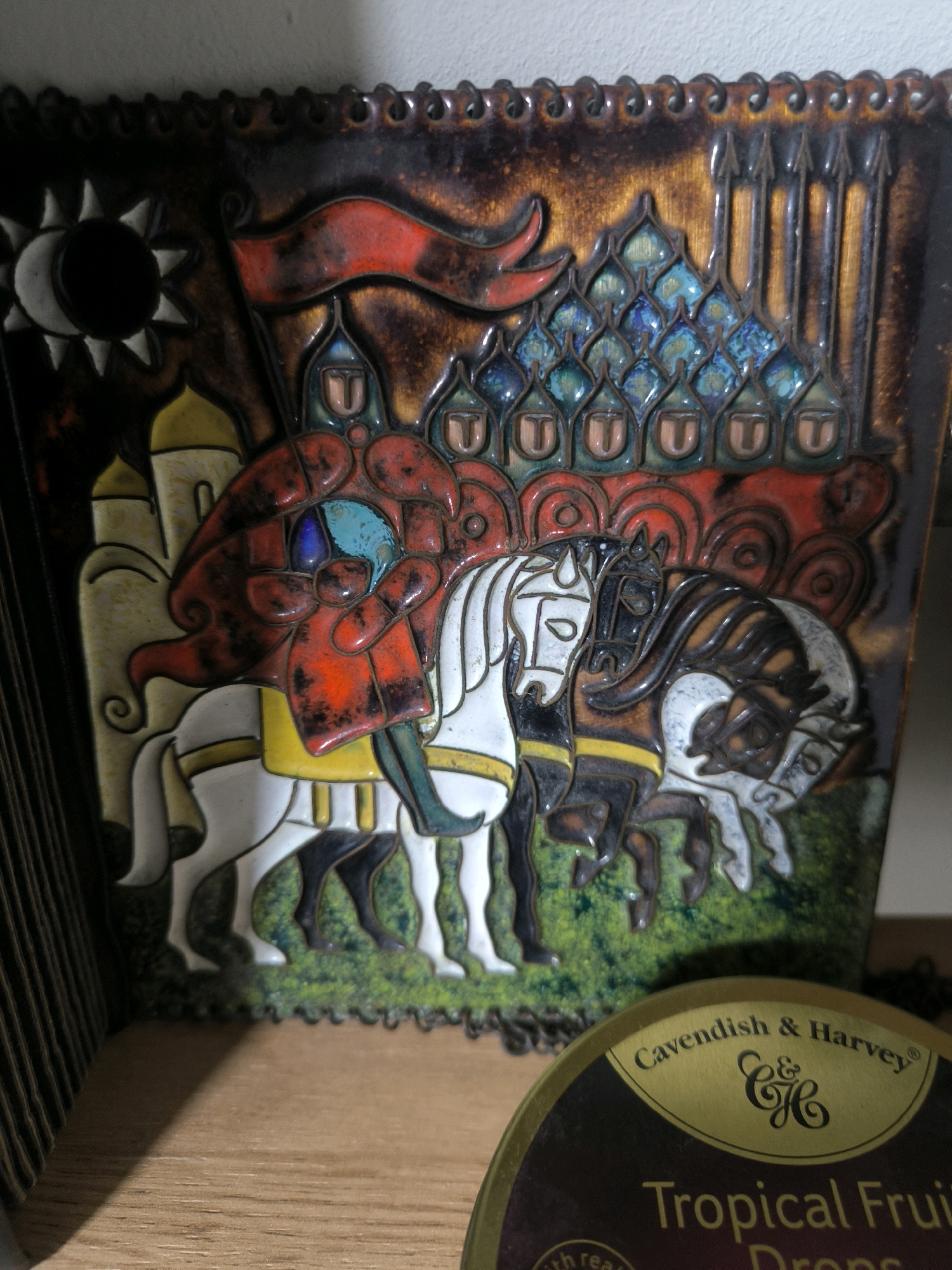 The image shows a close-up of a decorative ceramic tile depicting a scene with multiple horses and riders, possibly in a historical or mythical context. The tile features intricate details and vibrant colors, with a predominantly brown, gold, red, and blue palette. In the foreground, a partially visible round tin with the label "Cavendish & Harvey Tropical Fruit Drops" is present. The background is a light-colored wall. The ceramic tile appears to be mounted or displayed on a shelf. The specific location within the city of Magnitogorsk, Russia is not discernible from the provided image.