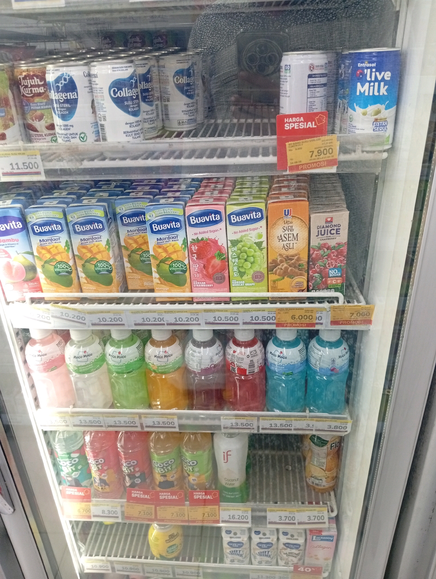 The image shows the interior of a refrigerated display case filled with various beverages in a store, likely a convenience store or minimarket, located in Bajingjowo, Indonesia. The display case is organized into shelves, each stocked with different types of drinks.
On the top shelf, there are cans of "Jujur Kurma" and "Collagen" drinks, as well as a blue can labeled "Enfatasol Live Milk." There is also a red promotional sign that reads "HARGA SPESIAL" (Special Price).
The second shelf from the top contains several boxed beverages, including "Buavita" juices in flavors like Jambu, Mangga, and Korean Strawberry, as well as "Ultra Sari Asem Asli" and "Diamond Juice Cranberry." Price tags are visible below these boxes.
The third shelf features bottled drinks, primarily "Mogu Mogu" in various flavors such as strawberry, grape, and orange.
The fourth shelf from the top holds bottles of "Coco Bit" drinks in different colors and flavors, also with "HARGA SPESIAL" promotional signs.
The bottom shelf shows more boxed drinks, including "if Coconut Water," a bottle of "FIO" juice, and several smaller cartons of milk or plant-based drinks, one of which is "Lactasoy Collagen."
The overall environment is clean and well-lit, with the cold air of the refrigerator visible as a slight mist or condensation on the glass door. No people are visible in the image. The arrangement of products suggests a commercial retail setting.