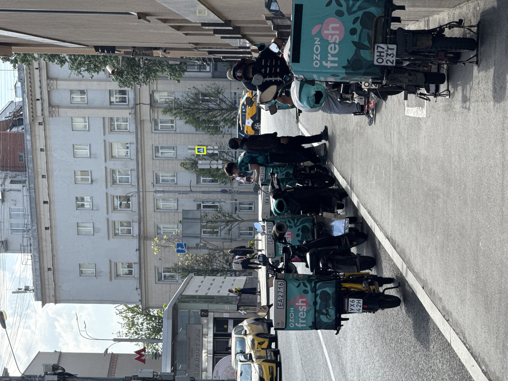 The image shows a city street in Moscow, Russia, lined with delivery scooters parked along the curb. The scooters bear the "OZON fresh" logo, suggesting they are used for grocery or food delivery services. Several people are near the scooters, presumably delivery drivers. The background features a large, multi-story building, typical of Moscow architecture, with numerous windows. Street signs and a Metro sign are also visible, indicating the location is near a transportation hub. The weather appears sunny, with clear skies.