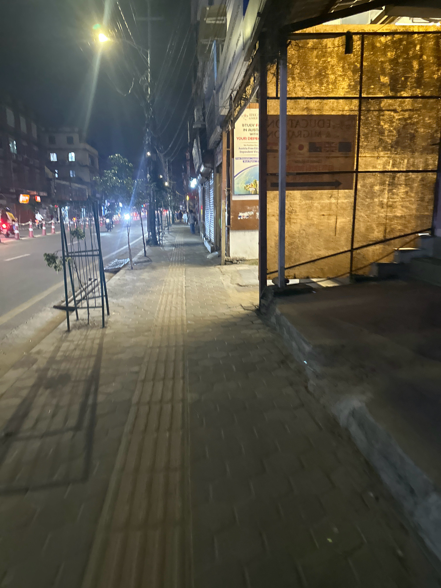 A nighttime street scene in Kathmandu Metropolitan City, Nepal, captures a pedestrian walkway alongside a road. The walkway is paved with textured tiles, some of which have raised tactile indicators, suggesting it's designed for accessibility. Along the edge of the walkway, a metal grate covers a drainage channel. Trees are planted in protective cages along the sidewalk, casting shadows from the artificial lights.
The road itself is asphalt, with visible lane markings and several traffic cones. A motorcycle is seen in the distance, illuminated by its headlights. The buildings lining the street are multistory, with illuminated windows and shopfronts. One building has a prominent yellow tarp covering its facade, behind which some signage is partially visible, including text that appears to be in English and related to education and migration services. The overall lighting is a mix of streetlights and building illumination, creating a moderate level of visibility. There are a few pedestrians visible on the sidewalk in the distance, walking.
