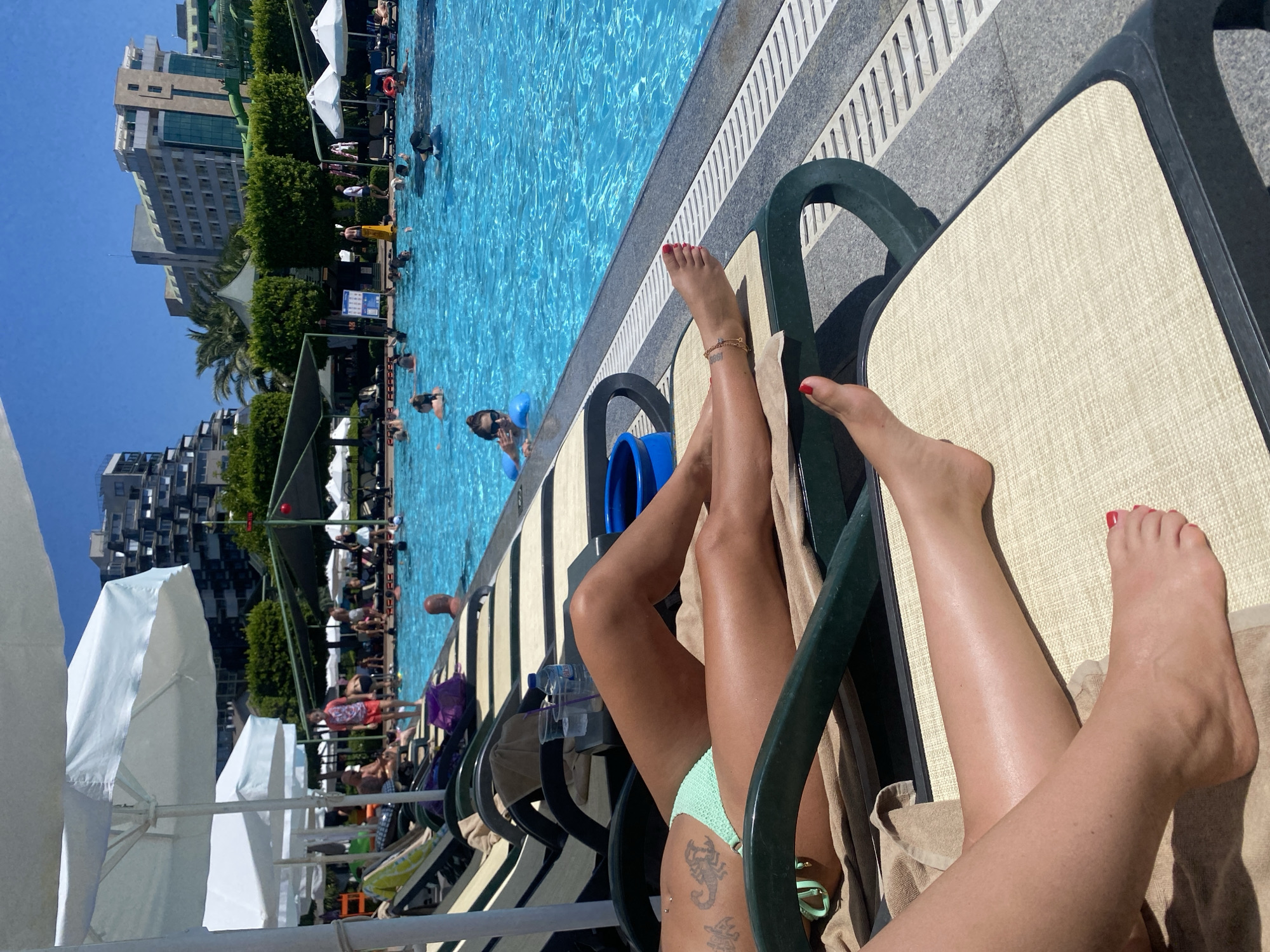 In Aksu, Turkey, at a resort, two individuals recline on lounge chairs by a vibrant blue swimming pool on a sunny day. One person's legs are visible in the foreground, adorned with a scorpion tattoo on their thigh. The other individual's feet are also visible, with red nail polish on their toes. Several other people are present in the pool, some swimming and others interacting. Large white umbrellas provide shade, and resort buildings are visible in the background. The scene depicts a leisurely vacation atmosphere.
