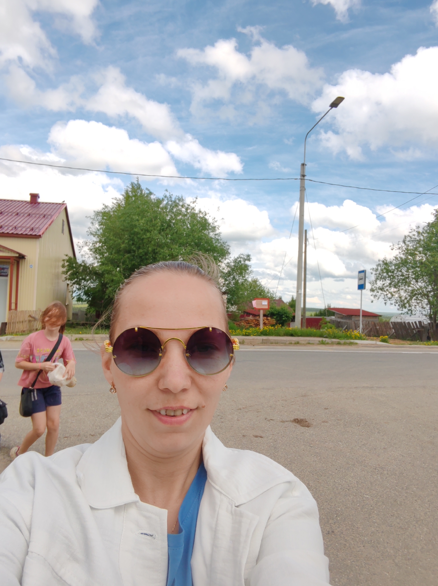 The image captures a selfie taken on a street in Чухлэм, Russia. A woman wearing aviator sunglasses, a white jacket, and a blue shirt is the primary focus. Behind her, on the left, a building with a red roof is visible, along with a younger individual walking away. The street includes a bus stop sign, a utility pole, and roadside vegetation. The sky above is partly cloudy.