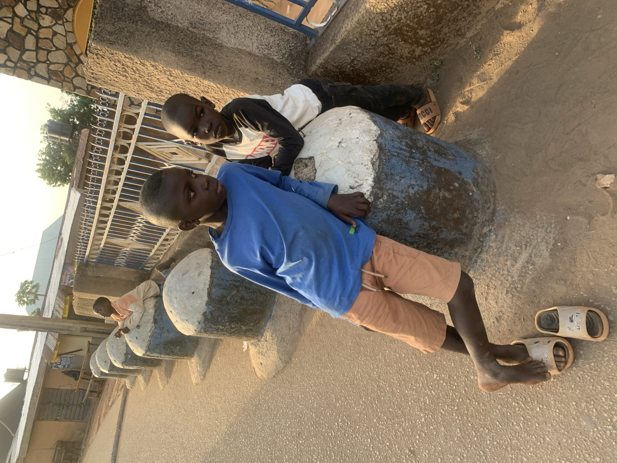 Two dark-skinned boys are lying on a series of concrete posts painted white and blue. The boys appear to be in their early teens. The boy closest to the camera is wearing a blue long-sleeved shirt and peach shorts. His feet are bare, with a pair of white sandals near his legs. The other boy wears a white long-sleeved shirt with black sleeves and black pants. He also wears brown sandals. They are both lying on the posts, with the concrete posts acting as seats. The background shows a street, buildings, and a fence with white railings, suggesting an urban area. The lighting indicates it is daytime. Given the presence of the concrete posts, street, and buildings, the scene likely depicts an everyday activity within a residential neighborhood. This image appears to be taken in Jos, Nigeria, due to various images of identical posts being widely reported in that city.