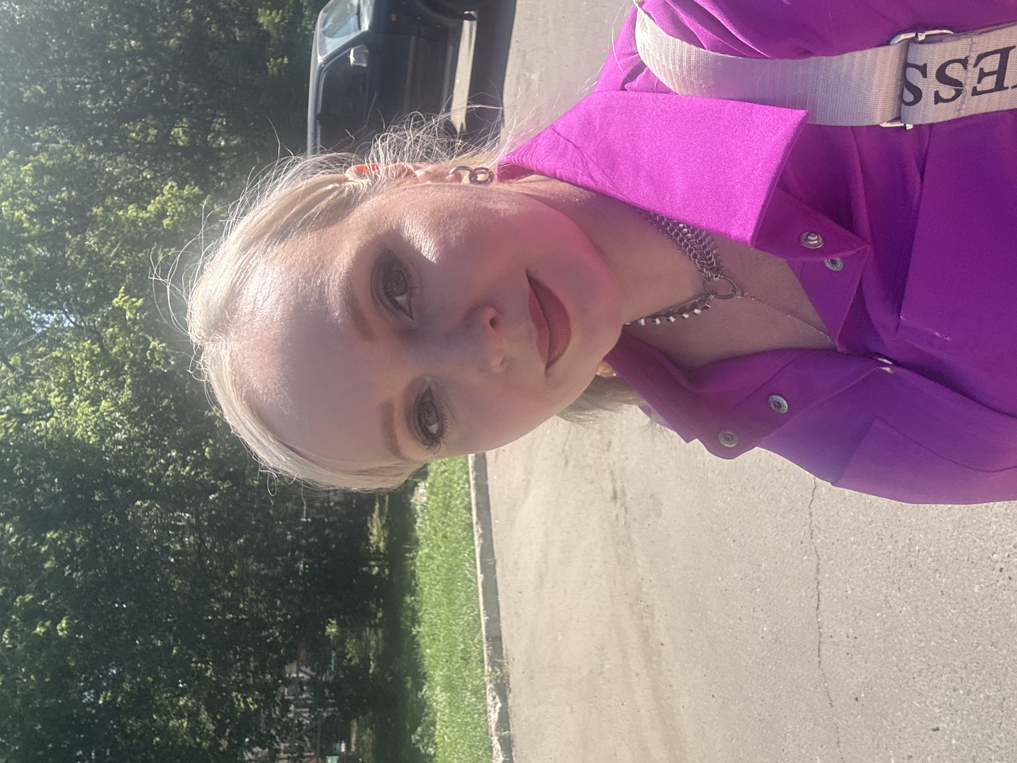 A woman with blonde hair and makeup takes a selfie outdoors on a sunny day. She is wearing a bright purple shirt and a silver necklace. A beige strap from a bag is visible across her shoulder, with the word "ESS" on it. Behind her, a dark-colored SUV is parked. The background consists of green trees and a paved area that appears to be a street or parking lot. The image was captured in Komsomolsk-on-Amur, Russia.