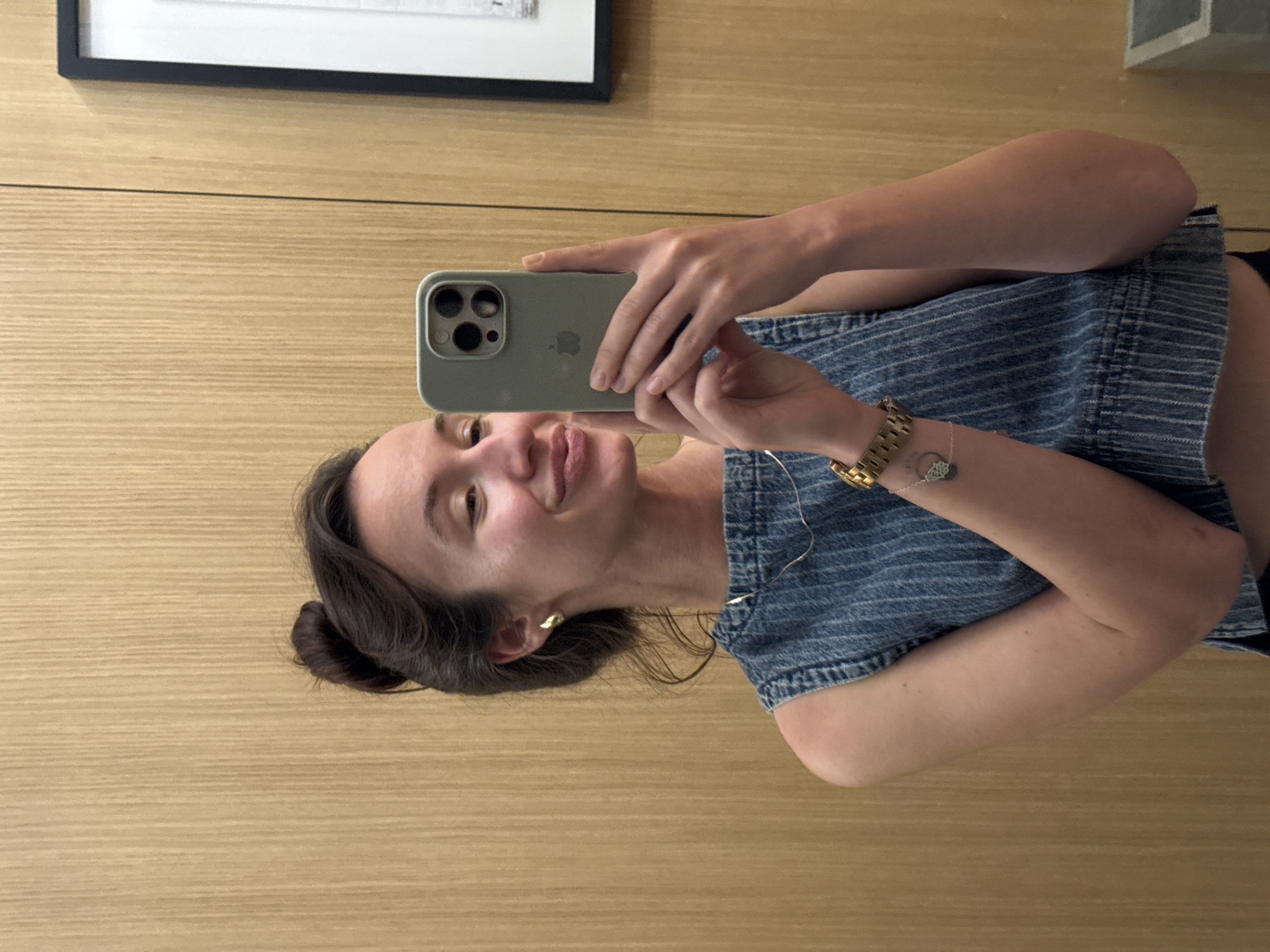 A young woman with her hair in a bun is taking a selfie in a room with wood-paneled walls. She is wearing a denim top and a gold watch. She is holding a light green iPhone in her right hand. A framed picture hangs on the wall behind her to the right. The scene appears to be indoors.