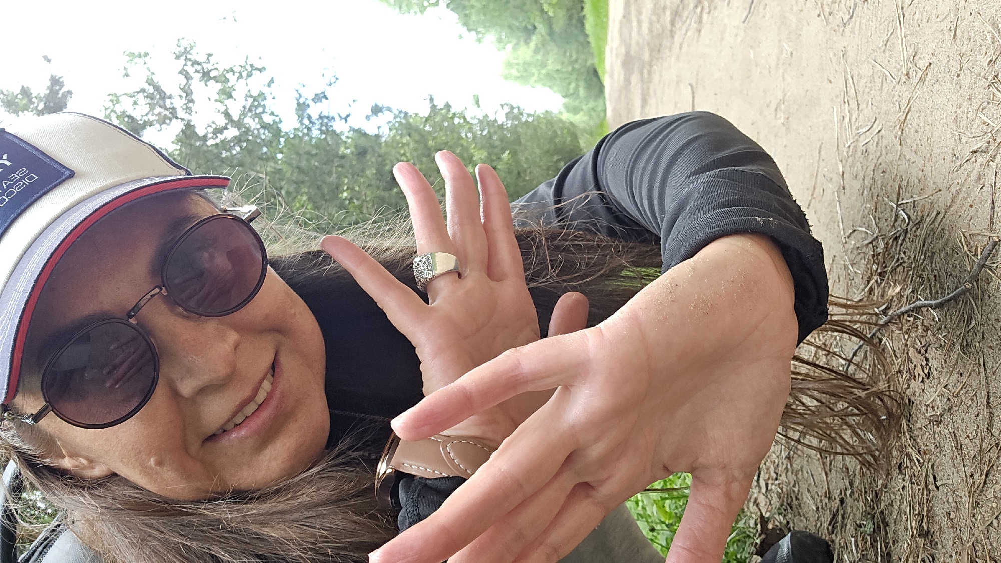A woman wearing sunglasses and a baseball cap smiles at the camera. Her hands are in the foreground, held up as if to block or shield herself. She is wearing a ring with a patterned band on her right ring finger. She is wearing a black long-sleeved shirt and appears to be outdoors in a natural setting with a dirt path visible behind her. The environment appears to be a wooded area. The image is a vertical, close-up shot.
