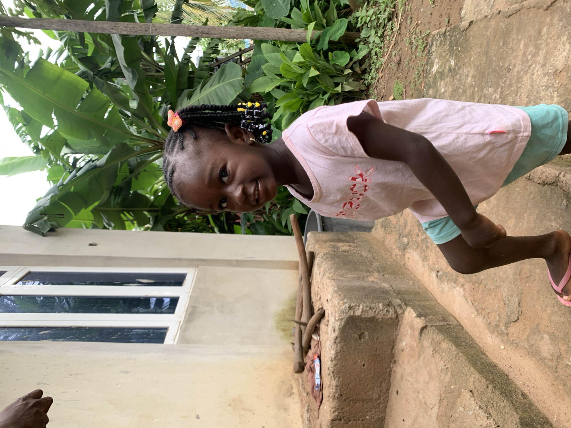 A young Black girl with dark skin and braided hair adorned with beads and a pink butterfly clip stands on a dirt path in front of a building with a light-colored stucco exterior and a white-framed window. She is wearing a light pink t-shirt and light blue shorts. The background consists of lush green banana trees and other foliage, suggesting a tropical environment. To the left of the girl, there are concrete steps with some wooden sticks and a piece of cloth lying on top. The overall setting appears to be a residential area in Akwanga, Nigeria, with the natural environment and the style of the building being characteristic of the region.