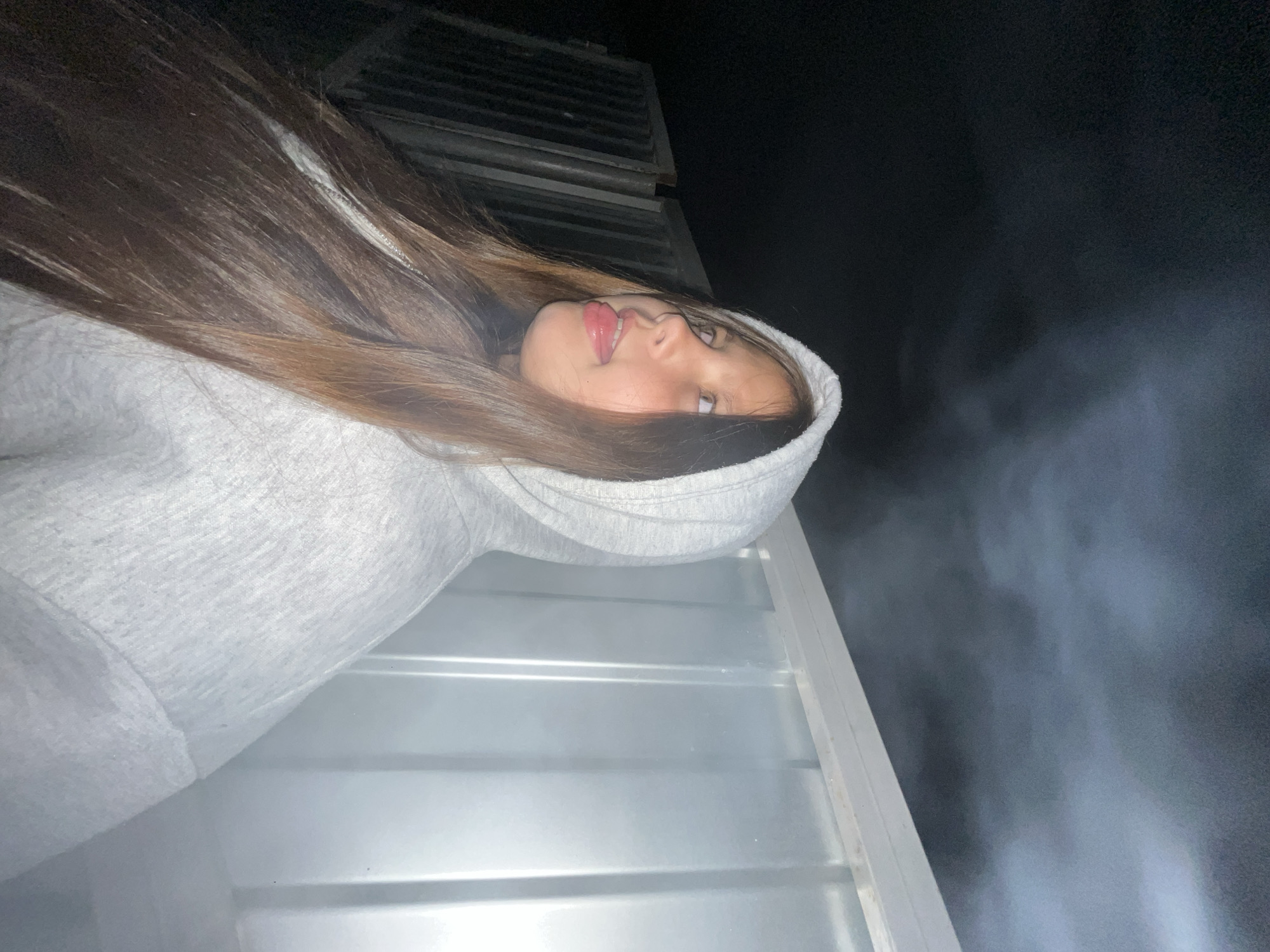 A young girl with long brown hair, wearing a light gray hooded sweatshirt, is looking to her left with her tongue sticking out. She is standing in front of a ribbed, white wall and to her left are dark, vertical structures that appear to be a fence or gate. The background is dark, suggesting nighttime. There is a misty or smoky effect in the upper portion of the image.