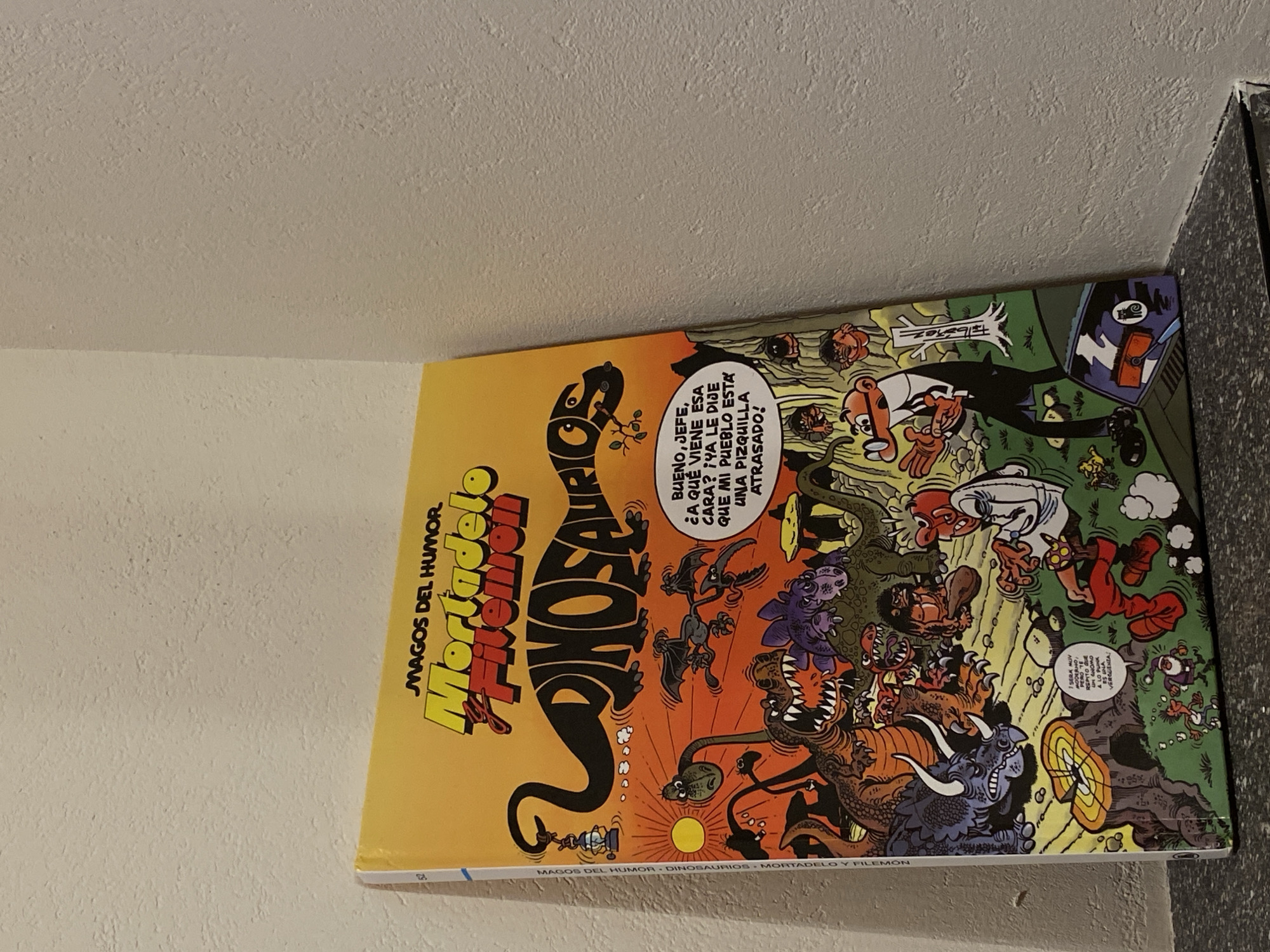 The image shows the cover of a comic book titled "Mortadelo y Filemón: Dinosaurios." The comic book is yellow with colorful illustrations depicting various dinosaurs and cartoon characters. The cover art features a large black dinosaur in the shape of the word "DINOSAURIO," surrounded by smaller dinosaurs, prehistoric human-like figures, and two main characters in suits. There is a speech bubble with Spanish text. The comic book is leaning against a textured, off-white wall. The bottom of the image shows a dark gray countertop with a black speckled pattern. There are no people visible in the scene, and the environment does not provide any specific clues about its location in Washington, United States of America.