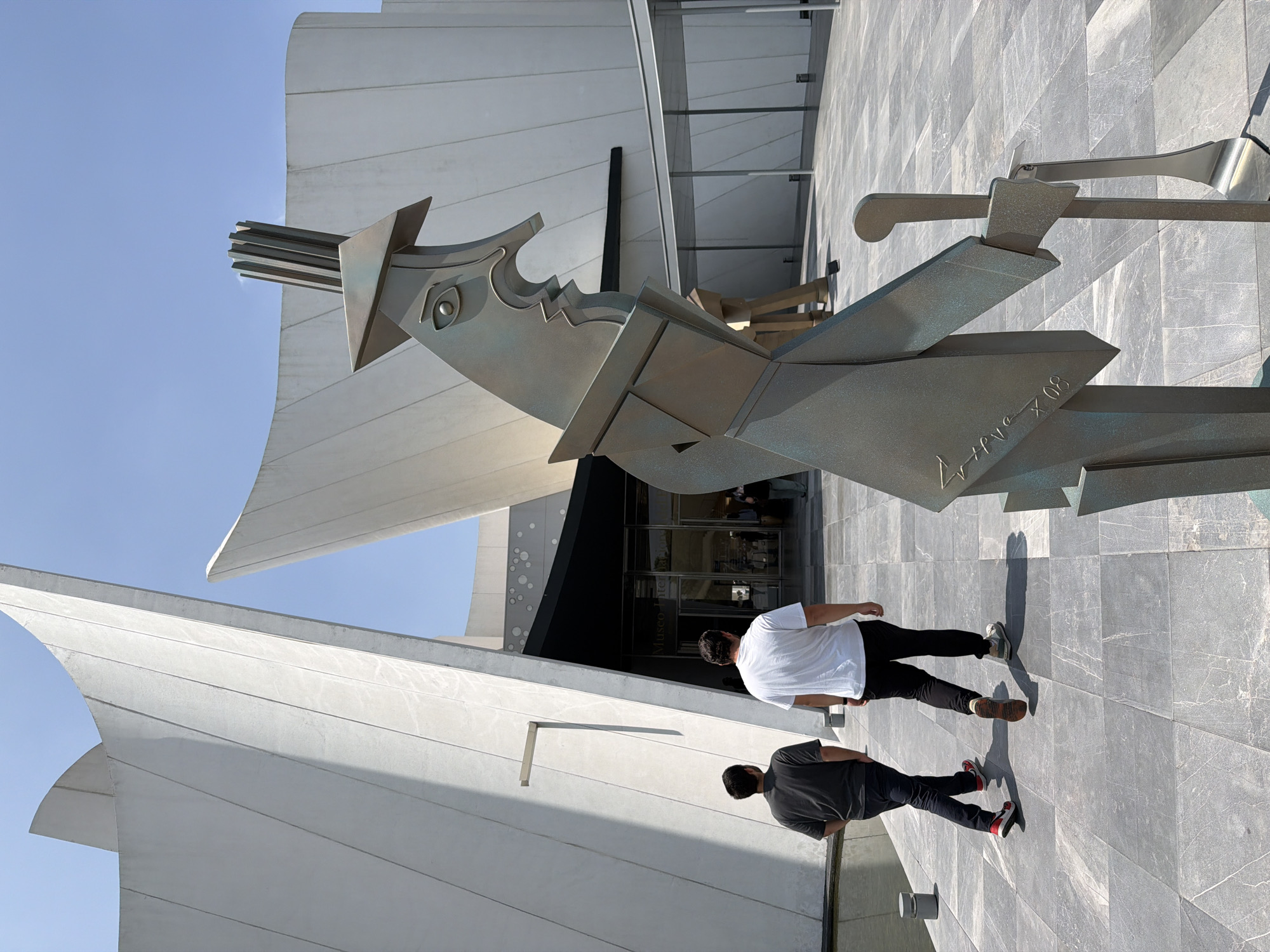 The image depicts a bronze sculpture in front of a modern building, with two men walking past. The building is white and angular, featuring large glass doors and a sign that reads "Museo Internacional." The sculpture is abstract, with geometric shapes and sharp edges, and appears to have an inscription on one side. The ground is tiled with gray stone. The sky is clear and blue. The men are dressed casually, one in a white shirt and black pants, and the other in a gray shirt and black pants. They are walking away from the camera, seemingly towards the entrance of the museum. This location can be found in San Bernardino Tlaxcalancingo, Mexico.