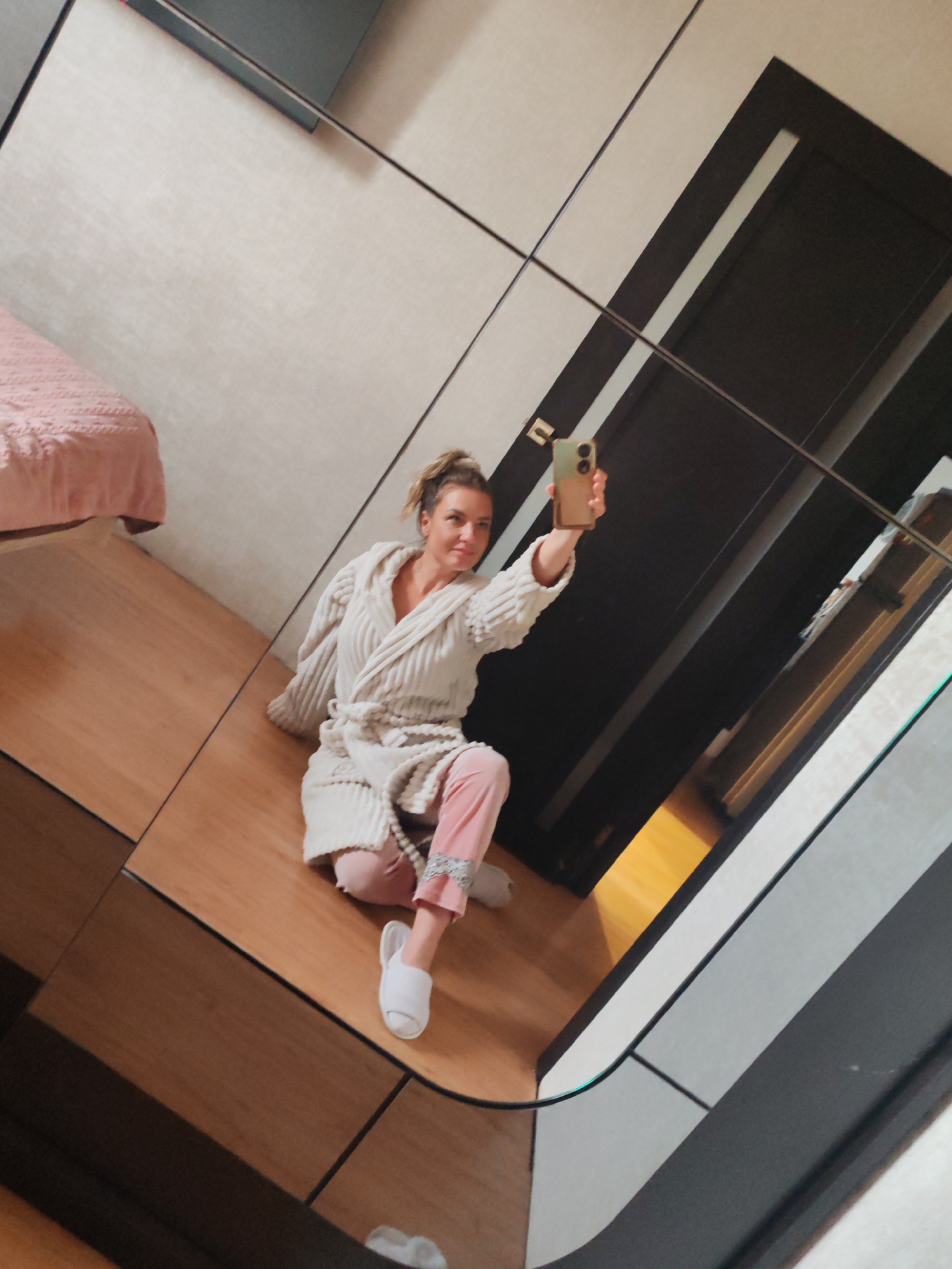 A woman is captured in a mirror selfie, seated on a light brown wooden floor, likely within a bedroom or dressing area in Petropavlovsk-Kamchatsky, Russia. She has light hair pulled back in a casual bun and is dressed in comfortable home attire: a plush, light beige textured bathrobe with a hood, pink pajama bottoms with lace trim at the ankles, and white slip-on slippers. Her legs are bent to the side as she sits, and her right arm is extended, holding a light green/beige smartphone to take the picture. She has a relaxed, pleasant expression, looking towards the phone.
The image is a reflection in a large mirrored surface, possibly a wardrobe or an accent wall, composed of multiple panels separated by thin black lines, which adds a geometric quality to the background. In the reflection, a bed with a light pink textured blanket is visible to the left. Behind the woman and to her right, a dark door or panel with a vertical light-colored strip is reflected. The room features light-colored walls and warm wooden flooring, suggesting a modern and tidy interior. The bright, even lighting indicates a well-lit indoor space, likely during the daytime. No visible text is present in the image.