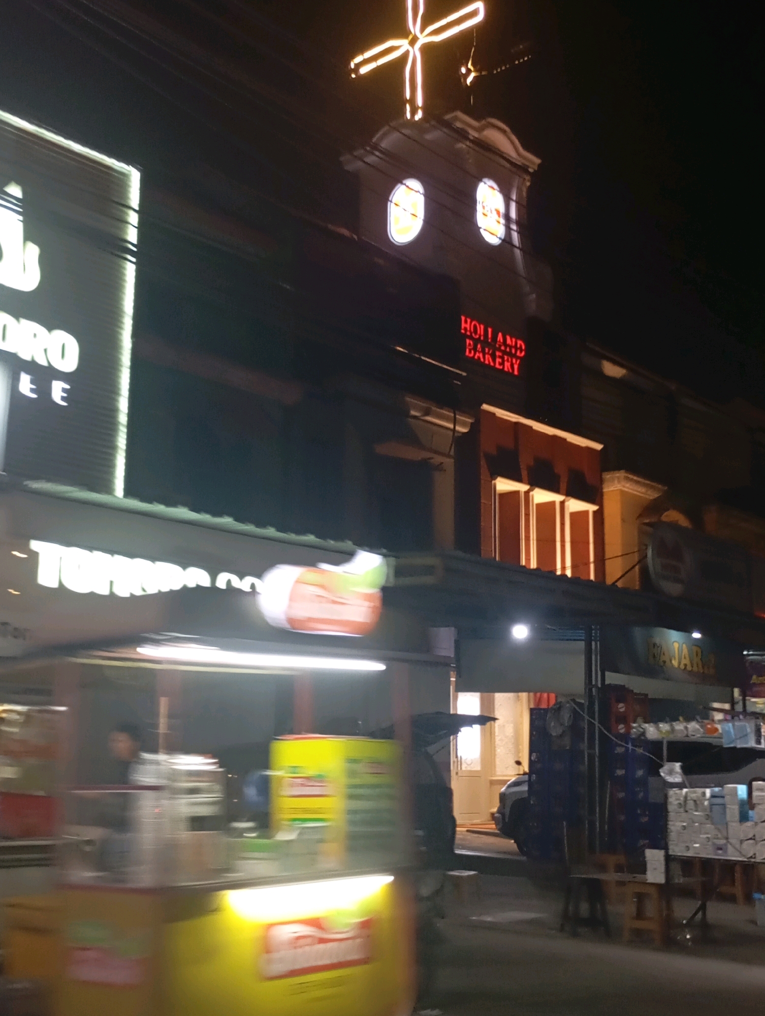 The image captures a nighttime street scene in Babelan, Indonesia. A prominent illuminated sign for "HOLLAND BAKERY" is visible on a building, accompanied by a large, cross-shaped neon light fixture above. In the foreground, a brightly lit food cart is in motion, creating a blurred effect. This cart displays various items and appears to have a person inside, partially obscured. Other businesses and structures line the street, some with their own illuminated signage, including one that reads "TOMOROCOFF." The overall environment suggests a commercial area at night with active street vendors and businesses.