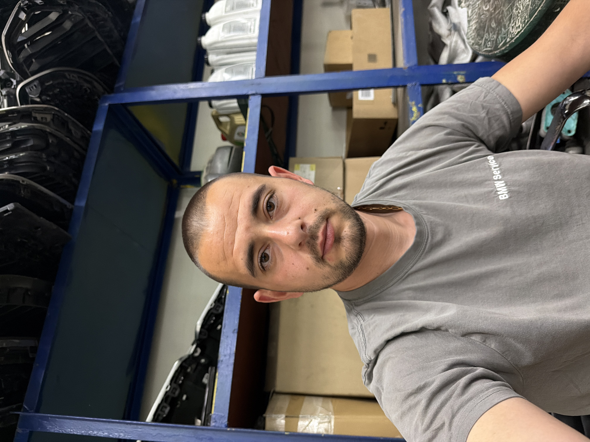 The image shows a medium shot of a man wearing a grey t-shirt with "BMW Service" printed on the left chest. He has short hair and a beard, and he is looking directly at the camera. The man is in what appears to be a garage or workshop, with shelves in the background holding various car parts and bottles of fluid. The shelves are blue metal, and some of the parts are car headlights. Boxes are stacked on shelves and on the floor. The lighting suggests an indoor environment, possibly illuminated by fluorescent lights. The overall impression is one of a professional automotive setting. The location is identified as Burgas, Bulgaria.
