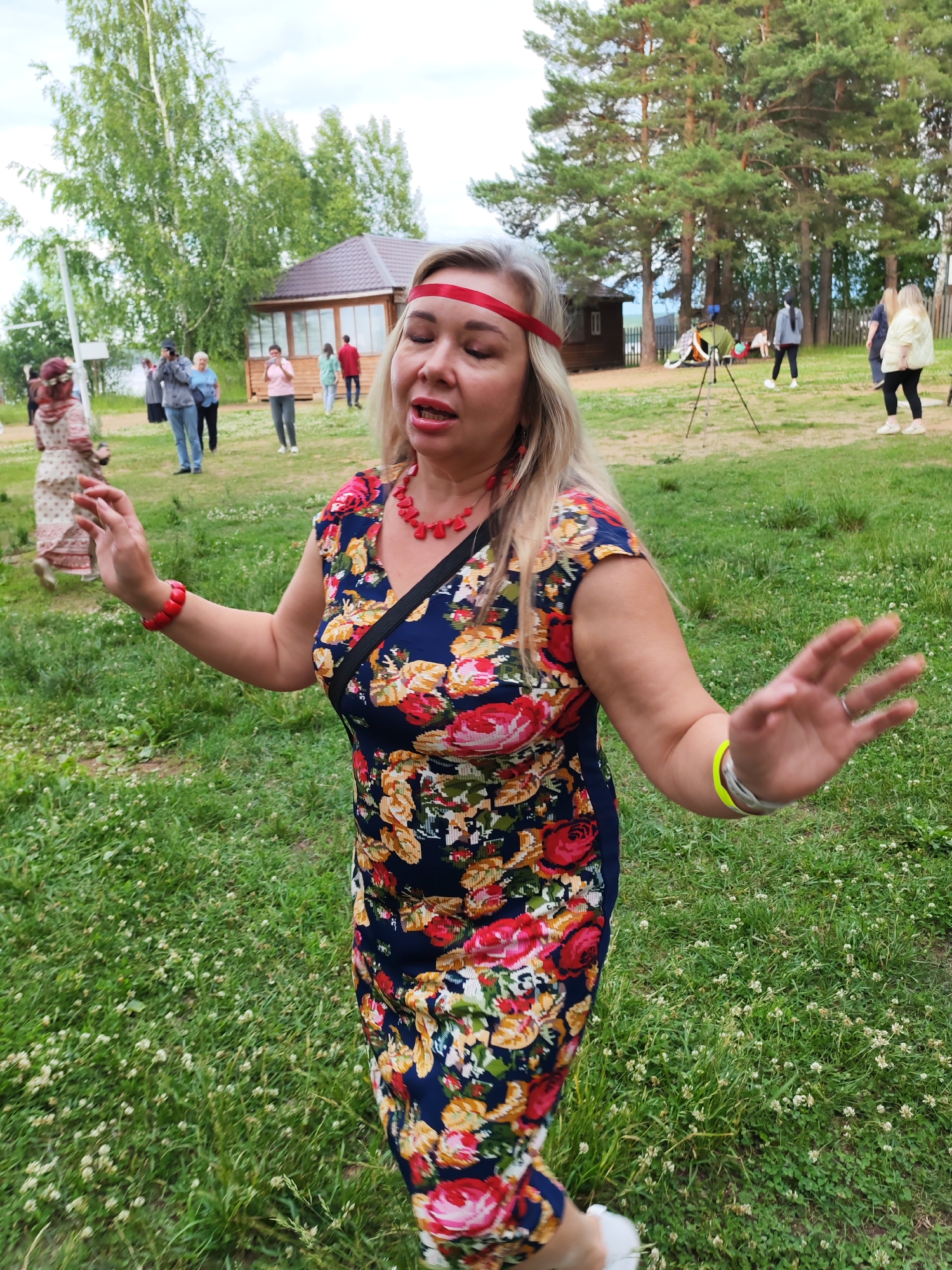 An outdoor daytime shot shows a woman in the foreground, wearing a floral dress and a red headband. She has blonde hair and is wearing a red necklace and bracelets. Her eyes are closed, and her mouth is open, suggesting she is singing or speaking. Her arms are outstretched, and her right hand is in a gesture of greeting or emphasis. In the background, other people are visible, some standing, some walking, near a wooden building and surrounded by trees. The setting appears to be a grassy area with some trees and a small wooden structure. The image was taken in the city of Chaikovsky, Russia.