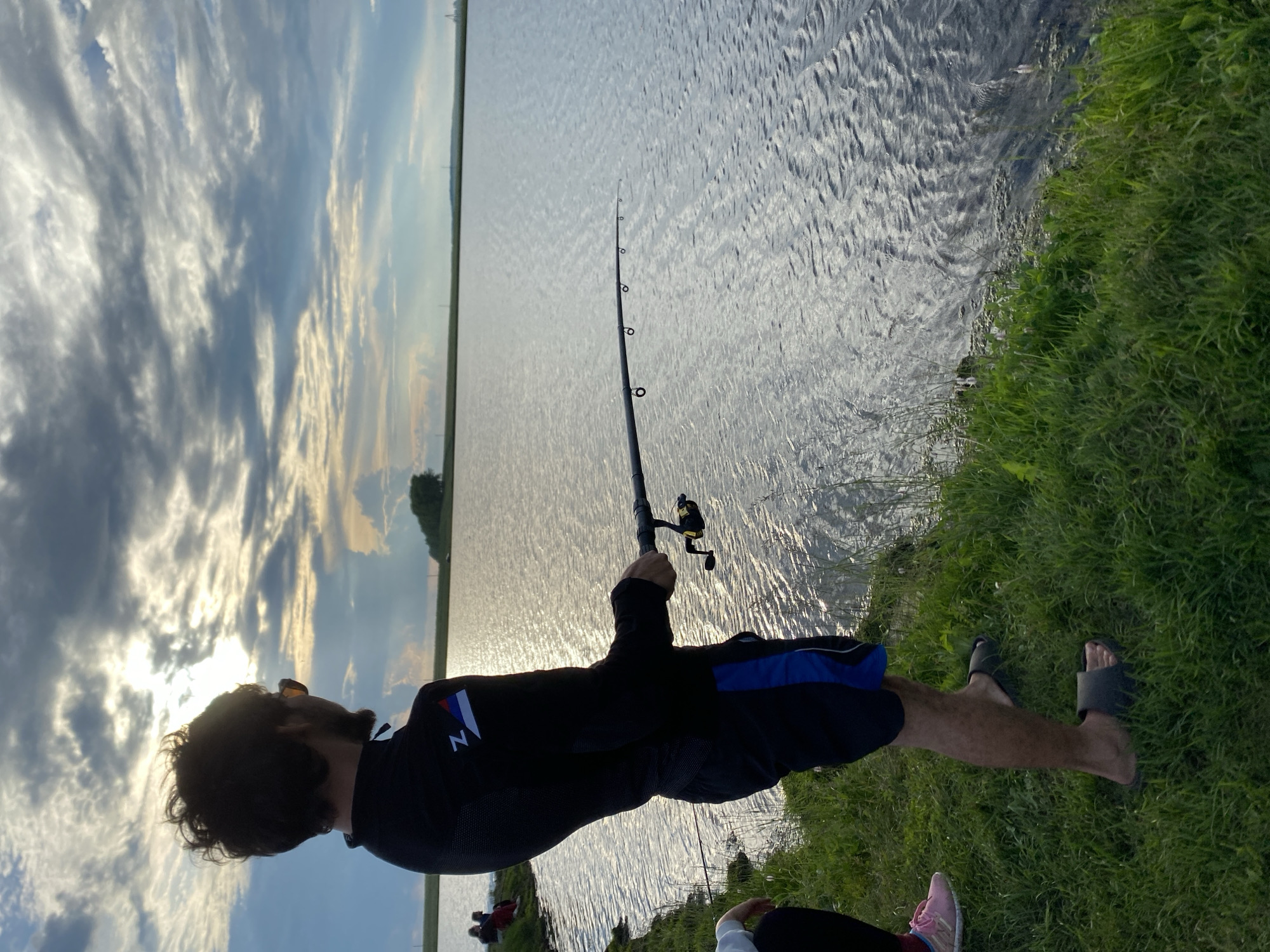 In the city of Сыйрышбашево, Russia, a person stands on the grassy bank of a body of water, fishing. The sun is partially obscured by clouds, casting a glinting reflection on the water's surface. The individual, facing away from the camera and towards the water, is wearing a black long-sleeved shirt with a logo on the left sleeve, black shorts with blue accents, and dark-colored slides. They are holding a fishing rod, actively engaged in fishing. To the left of the main subject, another person's legs are visible, wearing pink athletic shoes. The environment is natural, with lush green grass and a calm body of water stretching towards the horizon under a cloudy sky.