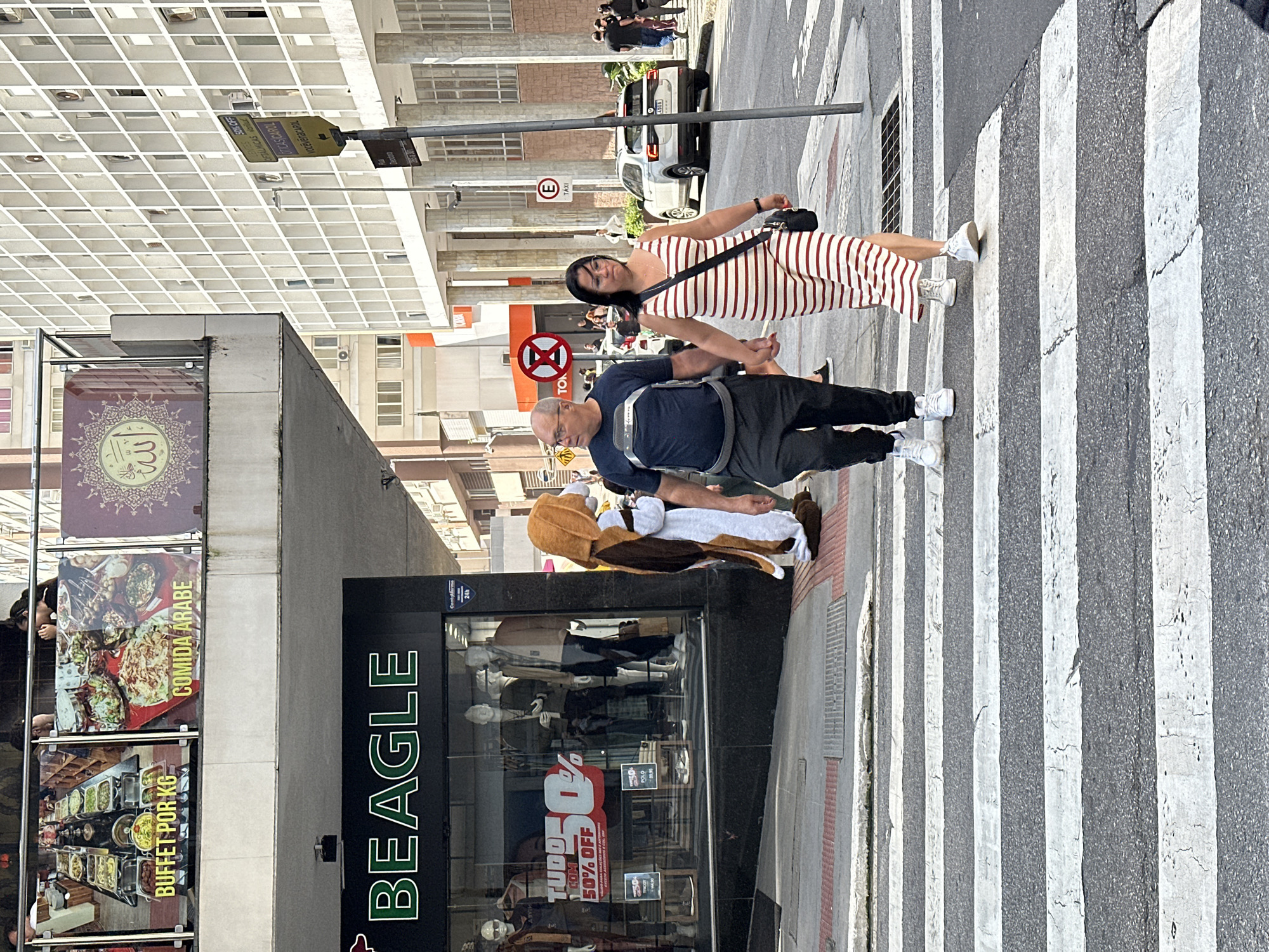 A full-shot photo captures a crosswalk in Florianópolis, Brazil, on a bright, sunny day. A couple, a man and a woman, walks hand-in-hand across the street. The man, wearing glasses, a dark blue t-shirt with a graphic design, and black pants, leads the way. The woman, with dark hair and glasses, wears a striped red and white maxi dress and carries a black crossbody bag. Behind them, a person in a beagle costume stands, partially obscured. In the background, a white car is parked on the right side of the street, and a modern, grid-patterned building stands tall on the right. A storefront on the left displays mannequins and sale signs, including one that reads "Beagle" in large green letters. The scene is a typical urban street in a Brazilian city.