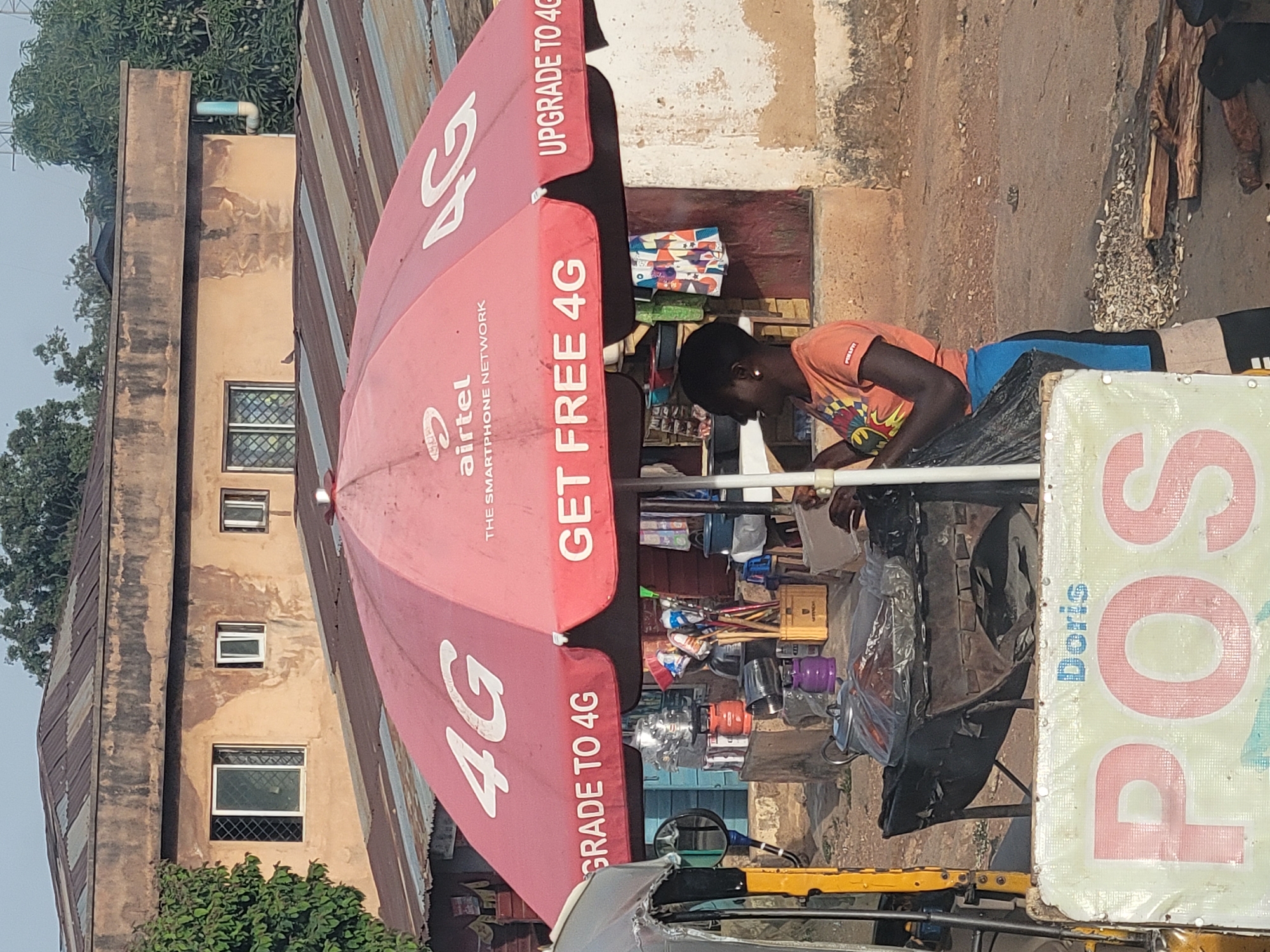 The image captures an outdoor street vendor stall in what appears to be a commercial area, consistent with a setting in Jos, Nigeria. A person, likely the vendor, is seated behind the stall, appearing to be engaged in an activity such as arranging goods or attending to a task. They wear an orange t-shirt with a graphic design.
A large, red advertising umbrella, prominently branded with "airtel THE SMARTPHONE NETWORK," shades the stall. The umbrella features large "4G" logos and slogans like "UPGRADE TO 4G" and "GET FREE 4G," suggesting the vendor offers telecommunication-related services or products.
The stall itself displays various items including small gas cylinders (one orange, one purple), what appears to be stationery, and other miscellaneous goods. To the right, a vertical sign prominently displays "POS" in large red and white letters, with "Doris" written in blue above it, indicating a Point of Sale service. A yellow part of a vehicle, possibly a tricycle (keke napep), is visible in the lower section of the image.
In the background, a multi-story building with faded, light brown walls and several windows fitted with security grilles is visible. The roof of this building and another nearby structure are made of corrugated iron. Green foliage and trees are present in the background. The scene is brightly illuminated by daylight, suggesting it is daytime. The overall impression is a vibrant, active street or market environment.