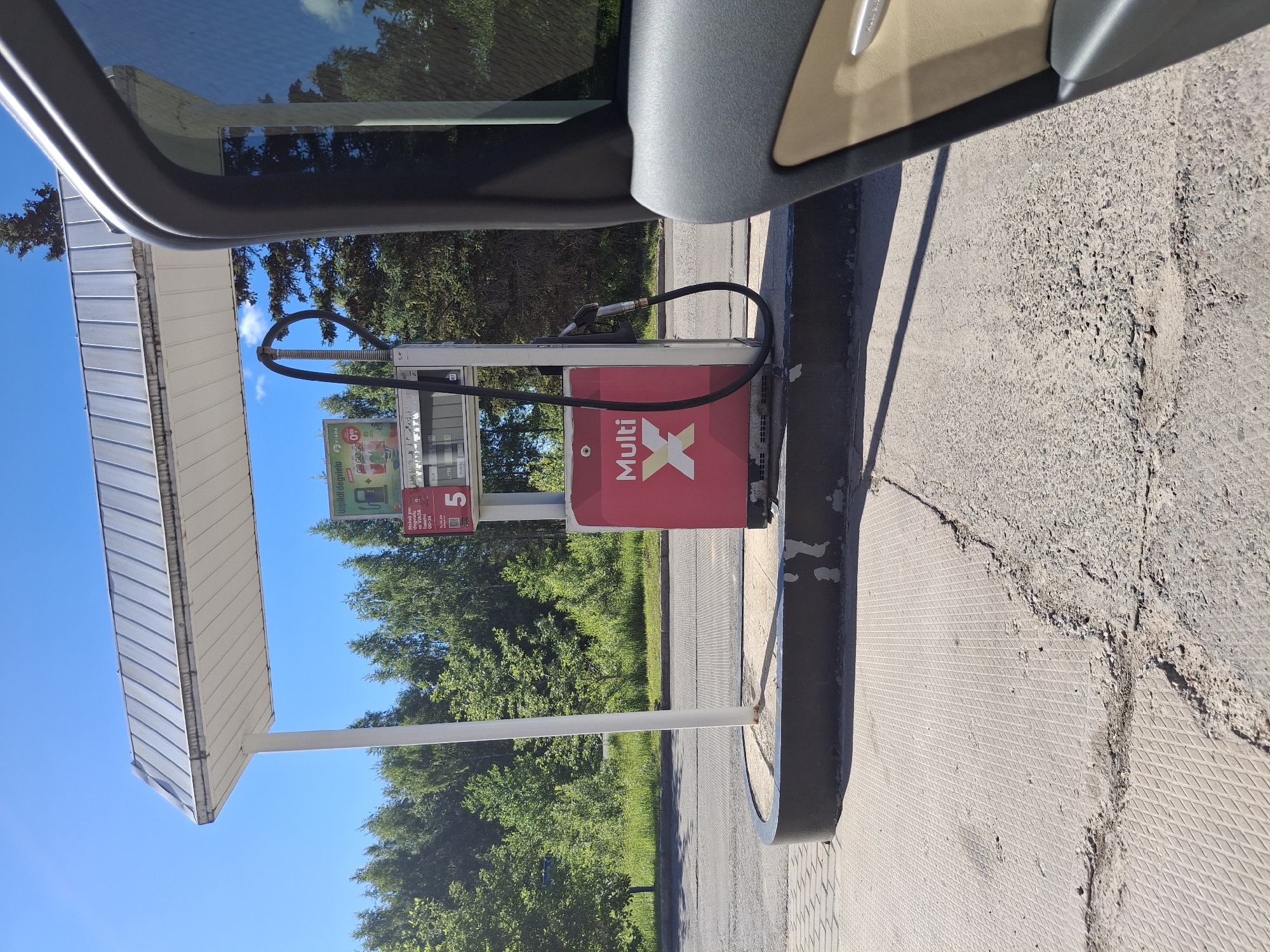 A gas pump at a station is depicted under a white awning in Glāžšķūnis, Latvia. The pump is red and white with the "Multi X" logo prominently displayed. A fuel hose hangs from the pump, ready for use. The surrounding area is paved with a textured concrete surface, which shows signs of wear and cracking. Green trees and lush vegetation form the background, under a clear blue sky. The right side of the image shows a partial view of a car, including its door and window, suggesting the photo was taken from inside a vehicle.