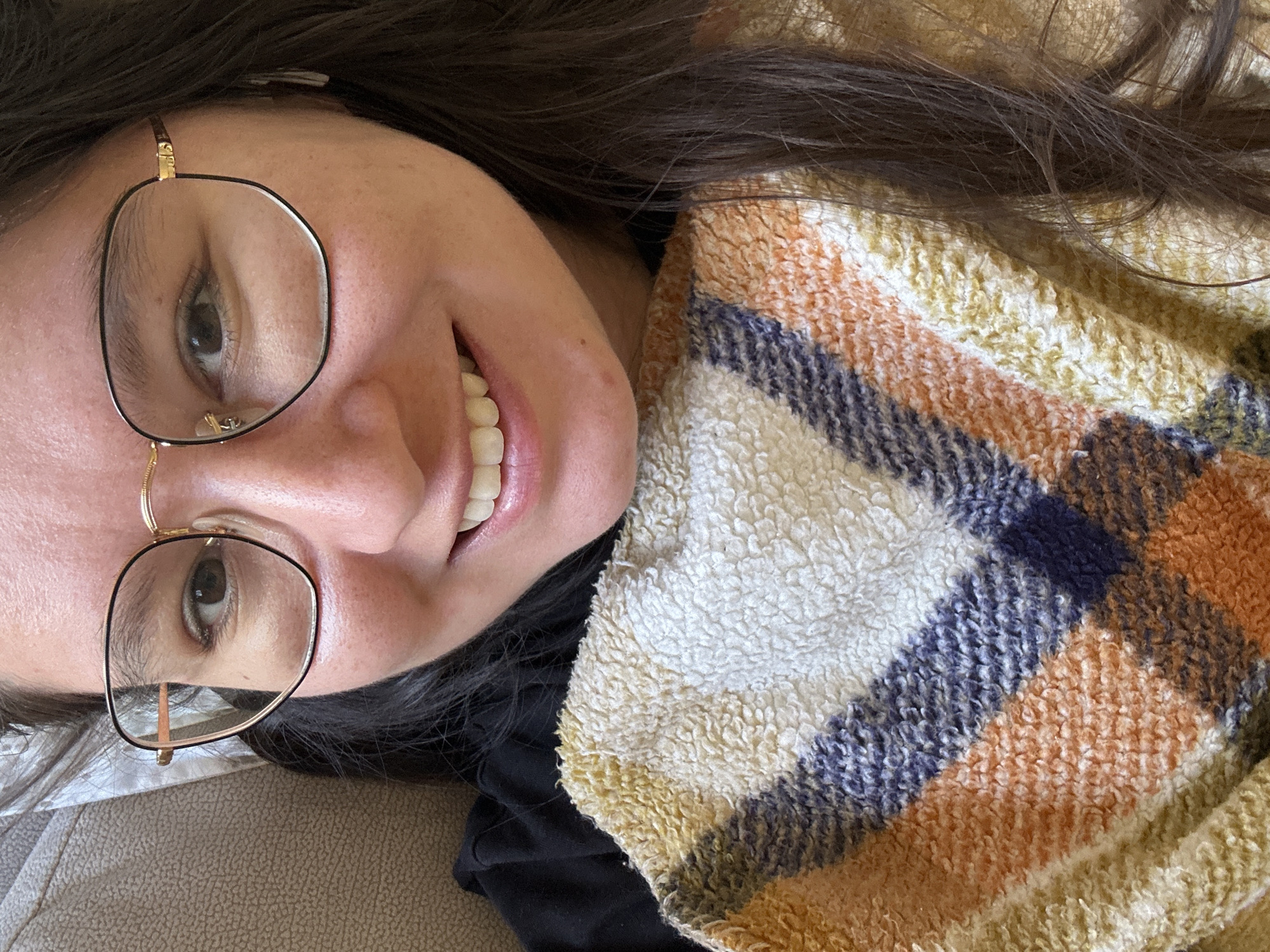 A woman is lying on a tan sofa, wearing glasses and smiling. She is partially covered by a plaid, fluffy blanket with colors of white, orange, navy, and yellow. She has dark hair. The location is not discernible.