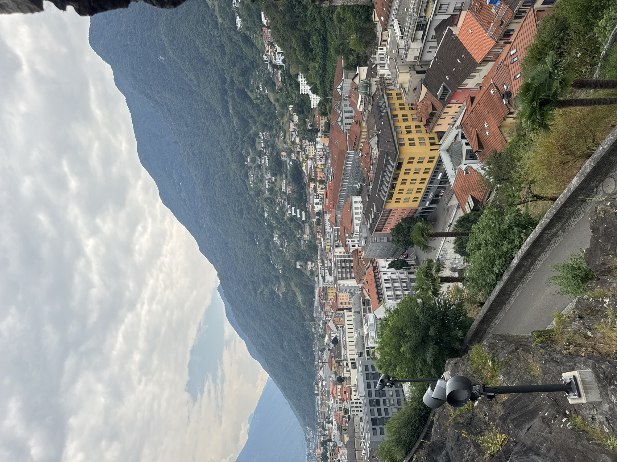 This is a wide, aerial shot of the city of Bellinzona, Switzerland, featuring a dense urban landscape nestled against a backdrop of lush green mountains. The sky is overcast with a heavy layer of clouds, creating a muted light. In the foreground, a rocky incline is visible with two modern, black floodlights mounted on metal poles, one of which is slightly out of focus. A paved pathway runs alongside the rocky edge, bordered by a low stone wall.
The city itself is characterized by a mix of architectural styles and colors. Buildings range from modern grey structures with numerous windows to older buildings with red tile roofs and brightly colored facades in yellow, pink, and orange. The urban sprawl extends up the mountainside, with houses and smaller structures dotting the green slopes. Several palm trees are interspersed throughout the scene, particularly in the lower right portion of the image, suggesting a temperate climate. The overall impression is of a city integrated with its natural mountainous environment under a cloudy sky.
