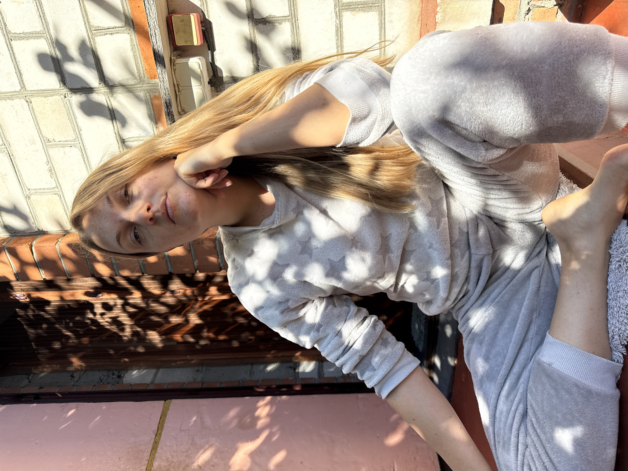 A young Caucasian woman with long blonde hair and freckles sits outdoors in front of a building. She is wearing a light gray plush pajama set with a star pattern on the top. Her left arm is bent, with her hand resting on her chin, and she is looking directly at the camera. The background consists of a brick wall with some electrical outlets and a wooden door, partially covered by the shadow of foliage. The scene is illuminated by sunlight, casting dappled shadows across the woman and her surroundings. This appears to be a residential area in Zheleznogorsk, Russia, based on the visual cues present.