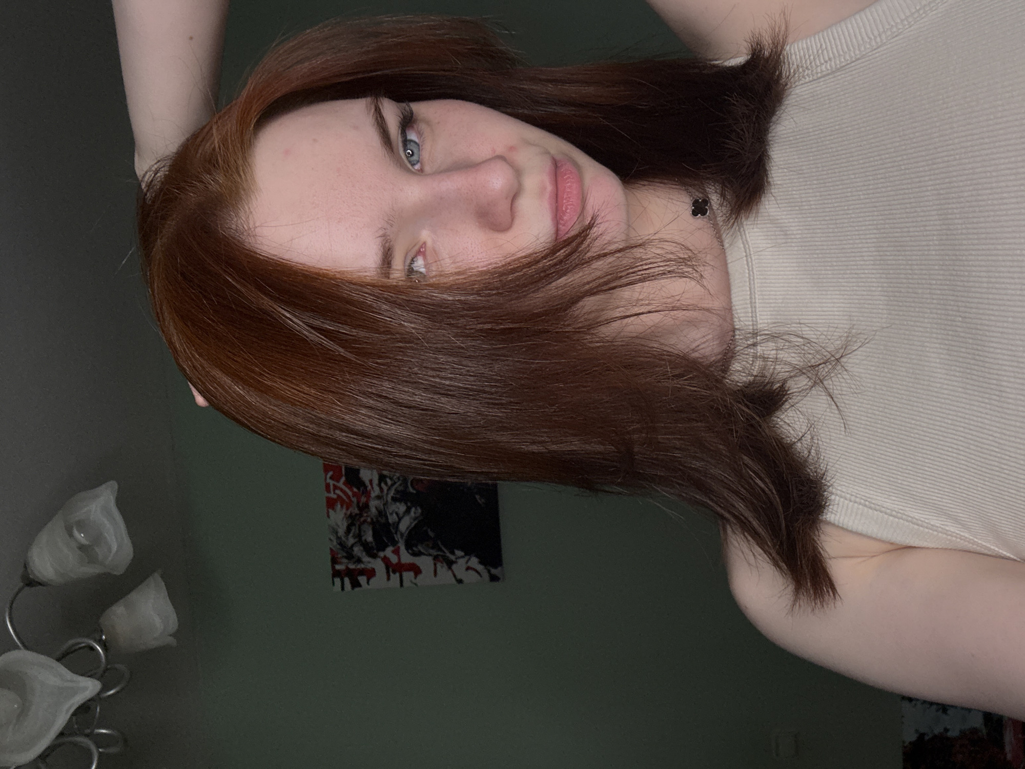 A woman with auburn hair and blue eyes is looking at the camera. She is wearing a light-colored ribbed tank top. Her right arm is raised above her head. Behind her is a green wall with a black and red abstract artwork. To the left is a lamp with three white tulip-shaped shades. The location is an interior space. The city is Unknown, Unknown.