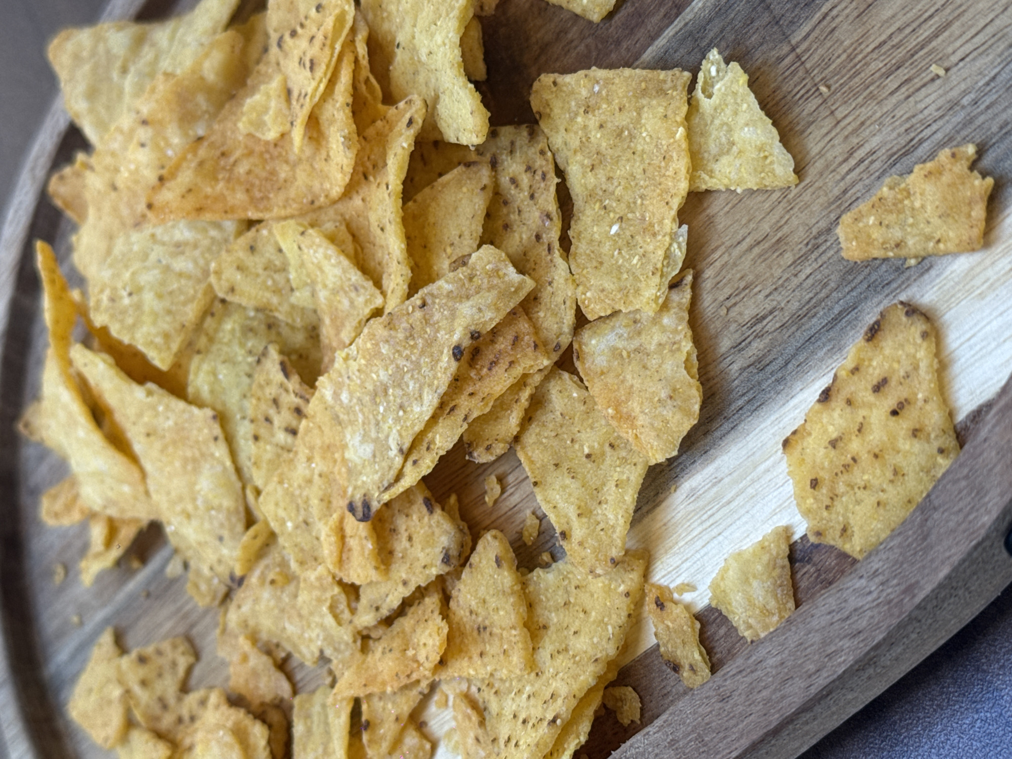 A close-up, top-down shot shows a pile of tortilla chips scattered across a wooden surface. The chips are triangular and have a golden-brown color with visible seasoning. Some chips are broken into smaller pieces. The wooden surface has a visible grain pattern and appears to be a serving platter or table. The lighting is natural, casting soft shadows on the chips. The image is sharp and focused, highlighting the texture and detail of the chips. There are no people or other objects visible in the frame. The scene is clean and uncluttered, suggesting a casual snack setting.
The image does not provide any information or context to determine a specific location in Podgorica, Montenegro.