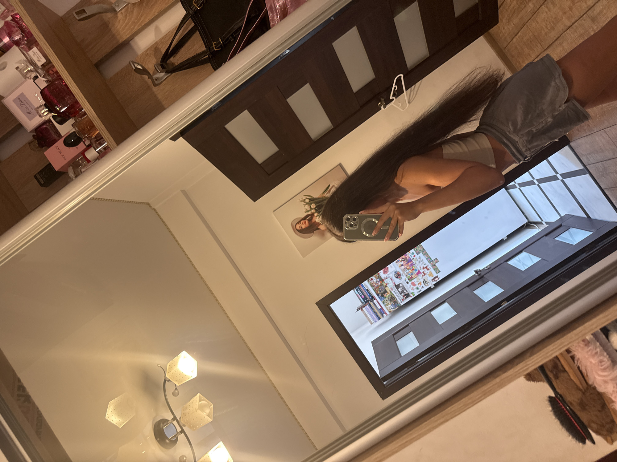 The image shows a young woman with long, dark hair, wearing a white crop top and gray shorts, taking a selfie in a mirror. She is holding a smartphone in her right hand, and her reflection is visible. Behind her, to the left, is a door with a frosted glass pane and a collage of images on the wall next to it. To the right of the door, there is a shelf filled with perfume bottles. A dark handbag hangs on a hook to the right. The room appears to be a bedroom or dressing area with a tiled floor and wooden doors. The overall lighting is dim, with light coming from ceiling fixtures and reflecting off the mirror. There is no visible indication of the location being in Chișinău, Moldova.
