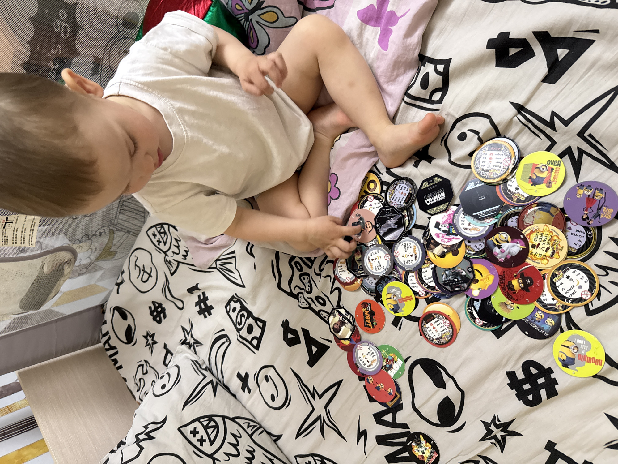 A young child, likely a toddler or preschooler, with light brown hair and wearing a white t-shirt, is depicted from an overhead perspective, engrossed in play. The child is situated on a bed or soft surface covered with patterned bedding featuring abstract black and white symbols, including dollar signs, stars, aliens, and various stylized characters, along with some pink and purple accents on a pillow.
The main activity involves the child playing with a large collection of round, disc-shaped collectibles, often referred to as pogs or milk caps. Many of these discs feature characters from the Minions franchise, while others display different animated characters, numbers, and descriptive words. The child is looking down at the discs, with one hand near the pile, suggesting focused engagement with the toys.
Notable details on the discs include various power stats like "FUN," "IQ," "POWER," "FAME," "Anger," "Love," "SPEED," and "ATTACK," often accompanied by numerical values. Several discs also show English phrases such as "OVER CONFIDENT," "I DON'T GIVE A PLUMOCK," and "WHAT DO YOU MEAN I'M EVIL." Cyrillic text is also visible on some discs, including "ЗВЕЗДНЫЕ ВОИНЫ" (Star Wars), "НИКО-КОЛИ," and "МАГАТ," strongly suggesting the location context of Rusanovo, Russia.
The setting appears to be an indoor child's play area or bedroom, partly enclosed by a mesh fabric wall, possibly a playpen or cot. A light wooden surface is visible in the foreground. The lighting suggests it is daytime, and the child appears calm and concentrated on their play.