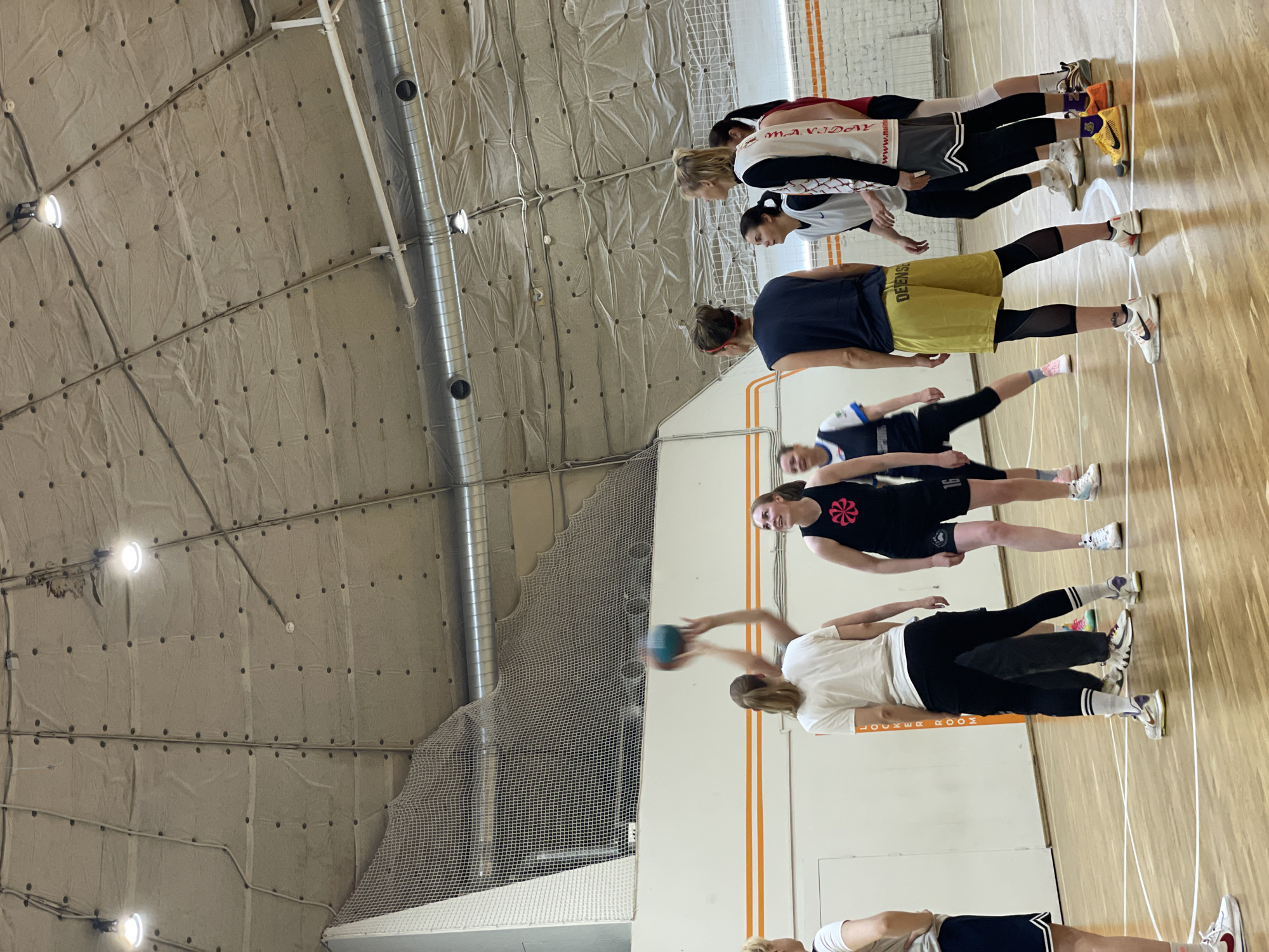 A group of women stand on a wooden court within an indoor sports facility. They are aligned in a row, and one woman is captured in the act of throwing a blue ball. The walls and ceiling of the facility are off-white and textured, with visible support beams and overhead lighting fixtures. The location is Saint Petersburg, Russia.