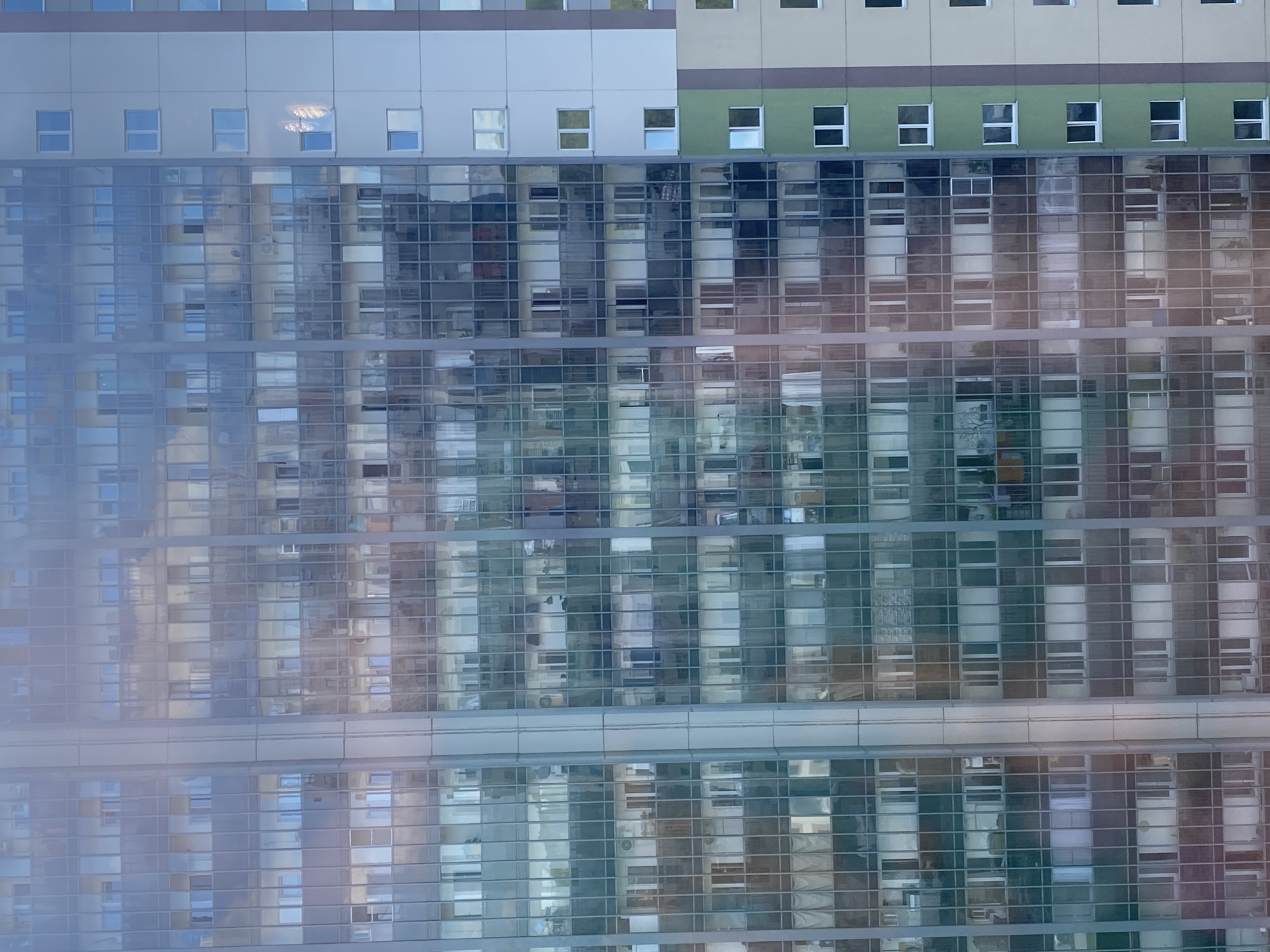 The image displays the facade of a multi-story apartment building, likely in the city of Novosibirsk, Russia, characterized by its modern architectural style featuring extensive glass and balconies. The building exhibits a grid-like pattern of windows and balconies, with some windows appearing illuminated, suggesting occupancy. Reflections on the glass facade indicate the presence of other buildings and possibly the sky. The building's exterior is a mix of glass, light-colored siding, and vertical sections of green and brown. The overall scene is static, with no visible human activity or movement. The environment appears to be urban, consistent with a residential area in a city.