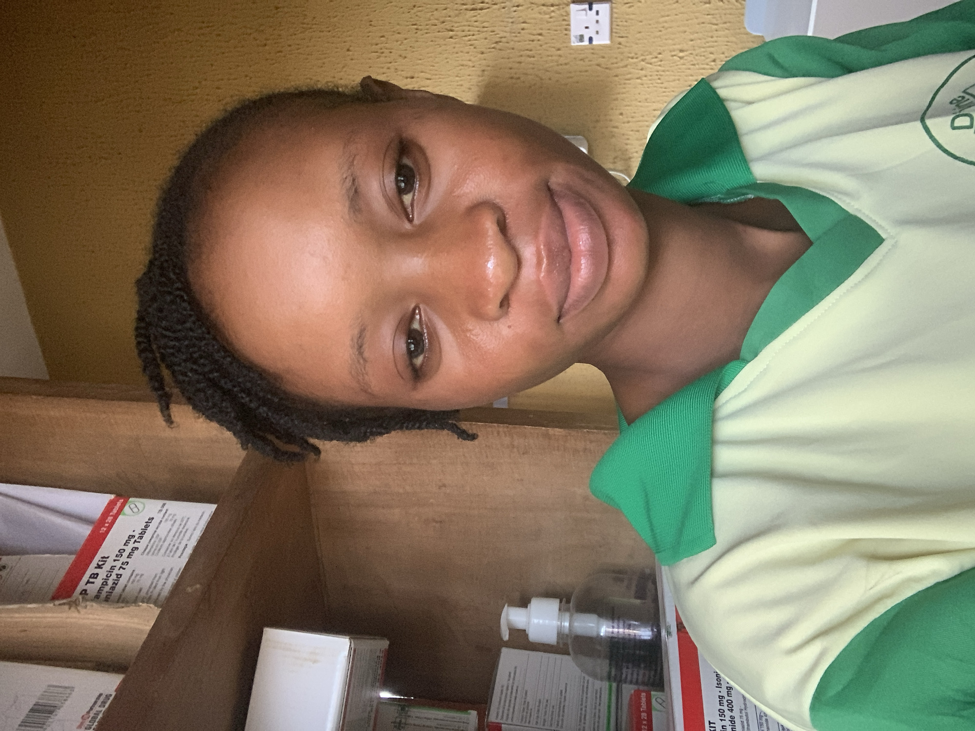 A selfie features a young Black woman in an indoor setting, likely a medical facility or pharmacy, given the surrounding objects. She has dark, braided hair pulled back, dark eyes, and full lips, looking directly at the camera with a slight, pleasant smile. Her skin appears well-hydrated. She is wearing a short-sleeved top that is primarily light yellow or cream-colored, with green accents on the collar, sleeves, and possibly side panels. A partially visible logo on her shirt includes the word "Dire."
The background consists of wooden shelves or cabinetry filled with medication boxes. Prominently visible is a box labeled "PTB Kit," indicating a kit for Pulmonary Tuberculosis, with text specifying "Rifampicin 150 mg," "Isoniazid 75 mg Tablets," and another box mentioning "Ethambutol Hydrochloride 400 mg." Several boxes bear the inscription "12 x 28 Tablets," and one box is marked "GLOBAL DRUG." Below the woman's face, a clear plastic hand sanitizer bottle with a white pump is visible on a shelf. The wall behind the shelves is textured and light brown or tan, and a white double electrical outlet is mounted on it in the upper right. The scene is likely located in Shagamu, Nigeria.