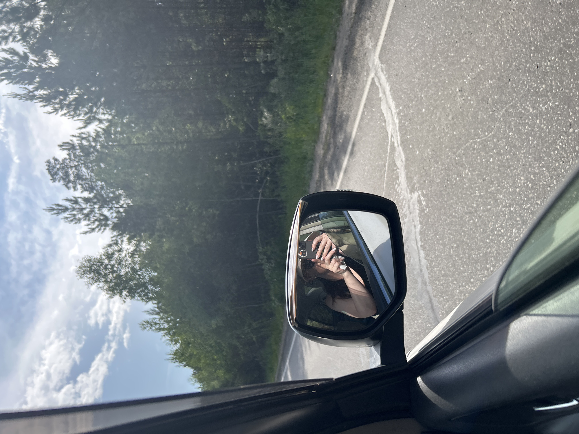 The image is a close-up shot from inside a car, looking outwards. The car is moving, as indicated by the reflection of the road and trees in the side-view mirror. A person is visible in the side-view mirror, taking a selfie with their phone. The scene outside the car is a road with trees and a cloudy sky. The sunlight suggests it is daytime. The overall mood is casual and relaxed, possibly indicating a road trip or a scenic drive.
The subjects of the image are:
- A person, seen in reflection in the side-view mirror. They are holding a phone and appear to be taking a selfie.
- The interior of a car, including the door frame and the side-view mirror.
- The exterior scene, which includes a road, trees, and a cloudy sky.
The activity taking place is a person taking a selfie while traveling in a car.
Notable details include:
- The reflection in the mirror shows the person's hands and face, as well as the phone they are using.
- The road appears to be paved and has some white markings.
- The trees are dense and green, suggesting a natural or forested area.
- The sky has scattered clouds.
- The time of day is likely daytime due to the bright lighting.
- The location is suggested to be Rodnik, Russia.
There is no visible text in the image.