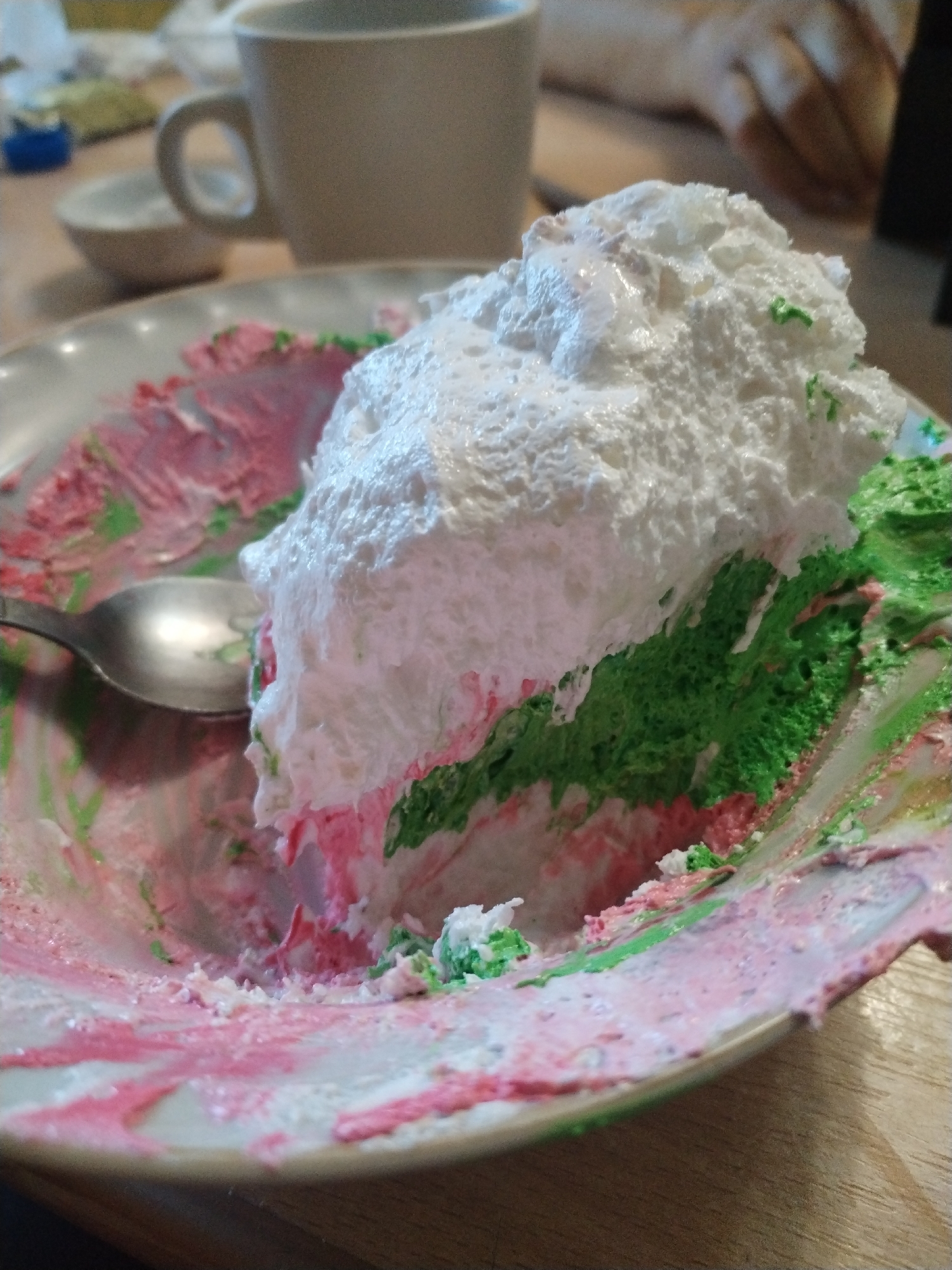 The image displays a close-up, eye-level, interior shot of a partially eaten, layered cake on a plate. The cake is topped with a large mound of white frosting and features visible layers of green and pink cake beneath. The plate is stained with pink and green frosting, indicating it has been served. A silver spoon rests on the plate to the left of the cake, partially submerged in the frosting remnants. In the background, out of focus, a beige ceramic mug and a small white dish are visible on a wooden surface. The lighting suggests an indoor setting with natural light. The scene is static, with no discernible human interaction depicted beyond the consumption of the cake. The location is unknown, and there are no identifiable city or regional markers.