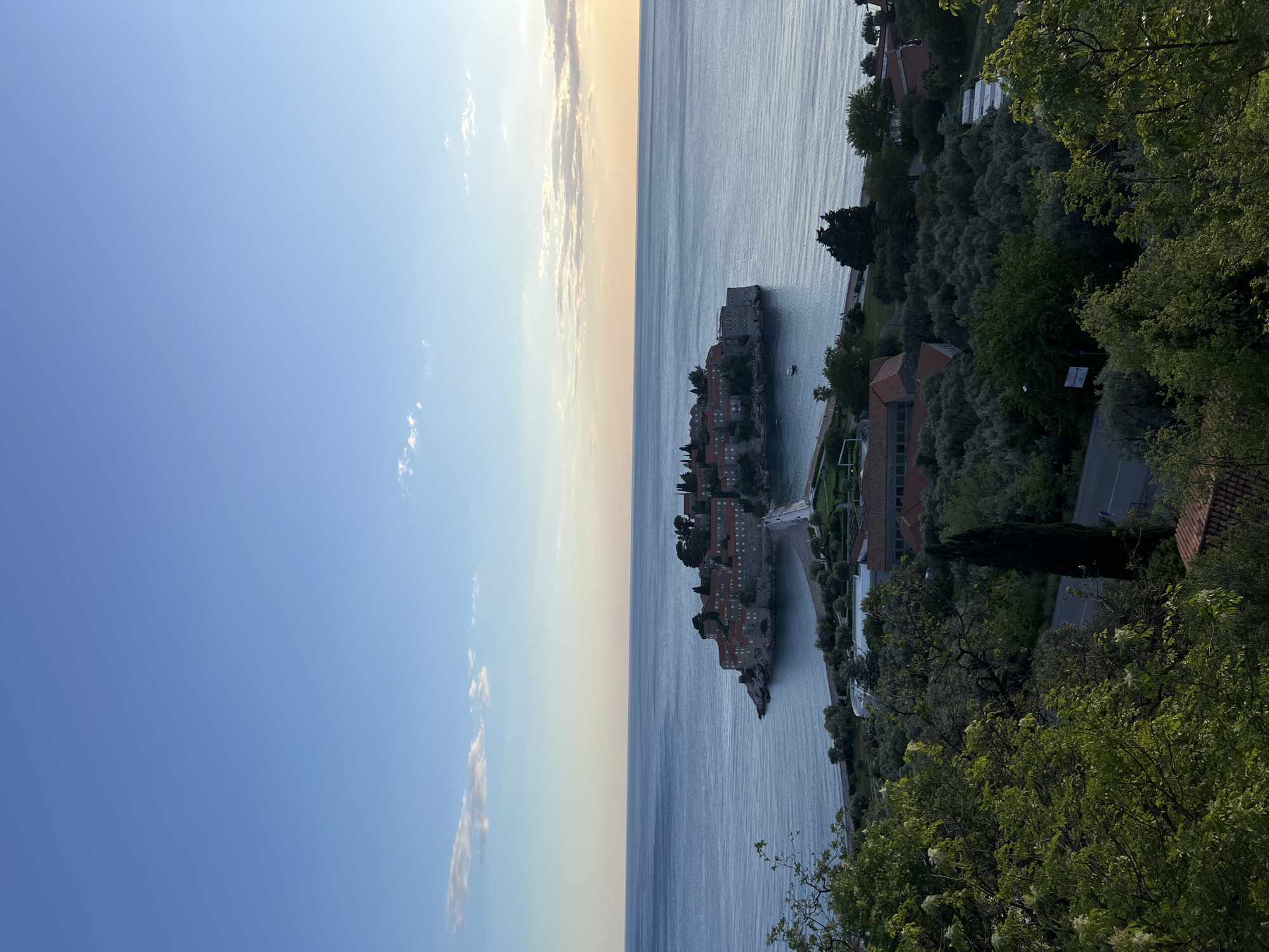The image presents a high-angle, panoramic view of Sveti Stefan, Montenegro, with the iconic islet taking center stage. The scene is dominated by the fortified village, a cluster of red-roofed buildings built on a rocky island, connected to the mainland by a narrow causeway.
The calm, deep blue waters of the Adriatic Sea stretch across the mid-ground and background, reflecting the soft ambient light. The sky, occupying the upper portion of the frame, displays a gradient from a clear, bright blue to a warmer, slightly golden hue near the horizon, suggesting the late afternoon or early evening hours, possibly around sunset. A few scattered, wispy white clouds are visible in the sky.
The foreground and the right side of the image (when viewed horizontally) are framed by lush green trees and dense foliage, indicating an elevated viewpoint overlooking the coast. On the mainland, alongside the causeway and further right, several buildings with red roofs are nestled within the greenery. No specific activity or people are discernible, contributing to the tranquil and picturesque nature of the landscape. Some small, unreadable signs appear on buildings on the mainland. The image evokes a sense of peace and natural beauty at a renowned Mediterranean destination.