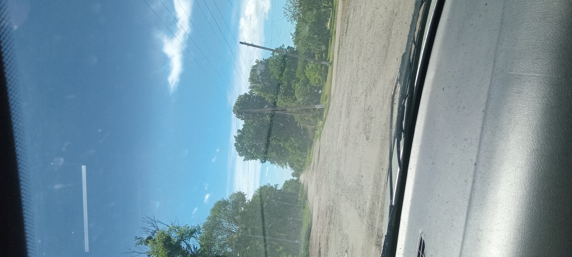 A point-of-view shot from inside a vehicle shows a dirt road lined with trees under a partly cloudy blue sky. The road is uneven and appears to be in disrepair, with visible potholes. Power lines are strung between wooden poles on either side of the road. The dashboard of the vehicle is visible in the foreground. The image appears to be taken in or around Koviahy, Ukraine, based on the context provided. There are no people or animals visible in the scene.