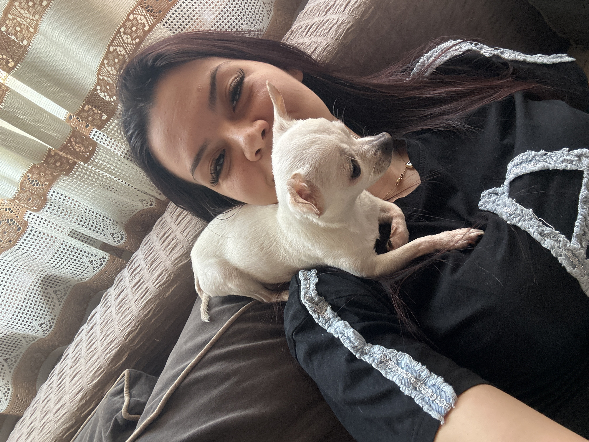 A light-skinned woman with dark hair is lying on a bed or couch, with a small, light-colored dog resting on her chest. The woman's head is tilted, and she is looking towards the camera with a slight smile. The dog is lying down with its head near the woman's neck. The woman is wearing a black top with light-colored decorative trim on the sleeves. A decorative, lacy curtain or fabric is visible in the background near the woman's head. The background appears to be a domestic interior. There is no information to indicate this image was taken in Skala, Bulgaria.