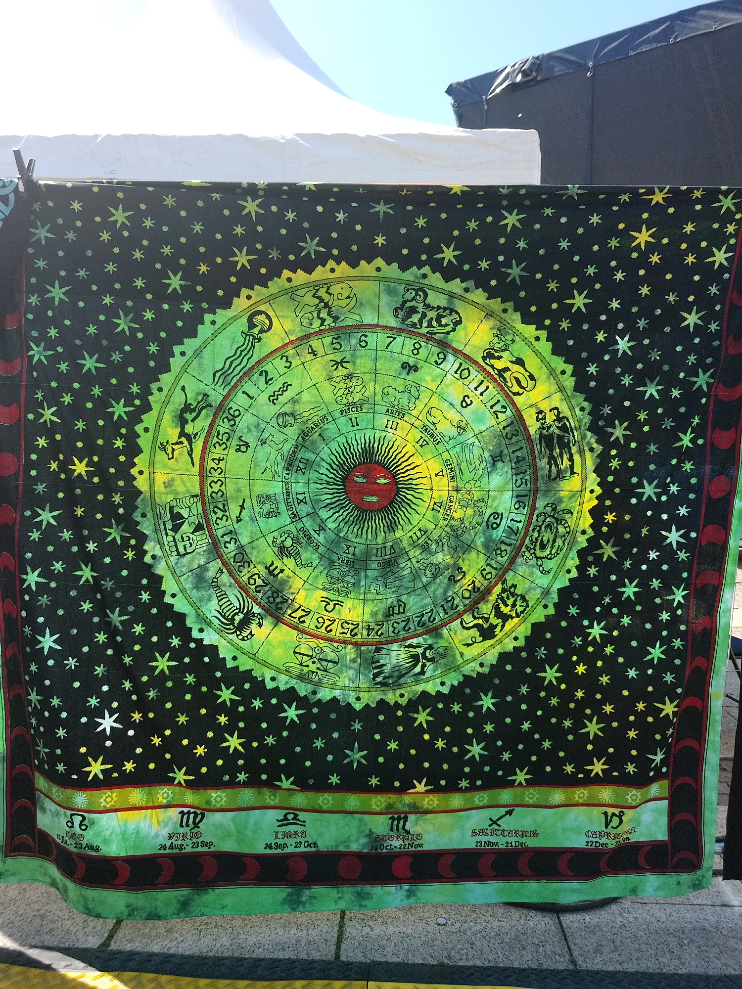 A tapestry displaying a zodiac wheel hangs from a makeshift clothesline outdoors. The backdrop of the tapestry is black and sprinkled with yellow and green stars. The zodiac wheel is rendered in shades of green and yellow, with a red sun at its center. Astrological symbols and zodiac signs are arranged around the perimeter of the wheel. Along the bottom edge of the tapestry, the zodiac symbols are displayed again with their corresponding dates. The backdrop includes a white canopy and a black structure, suggesting an outdoor market or fair. The ground appears to be paved with gray bricks or stone.